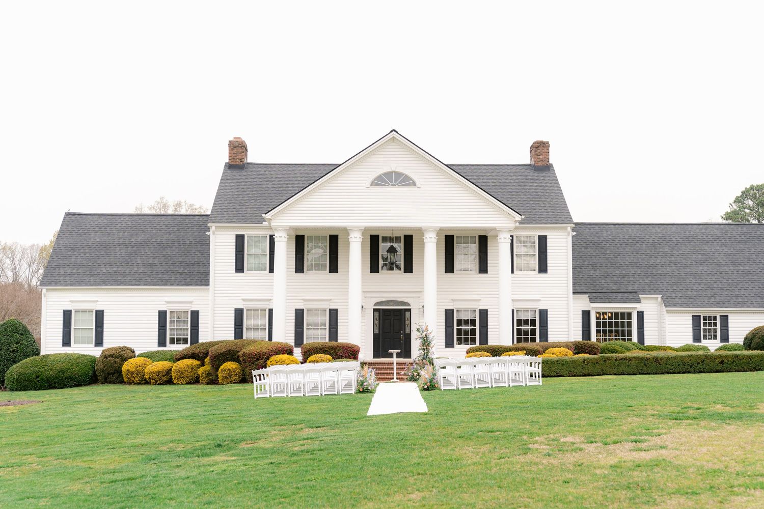 Elegant white colonial mansion with black shutters and yellow bushes sits on a manicured green lawn.
