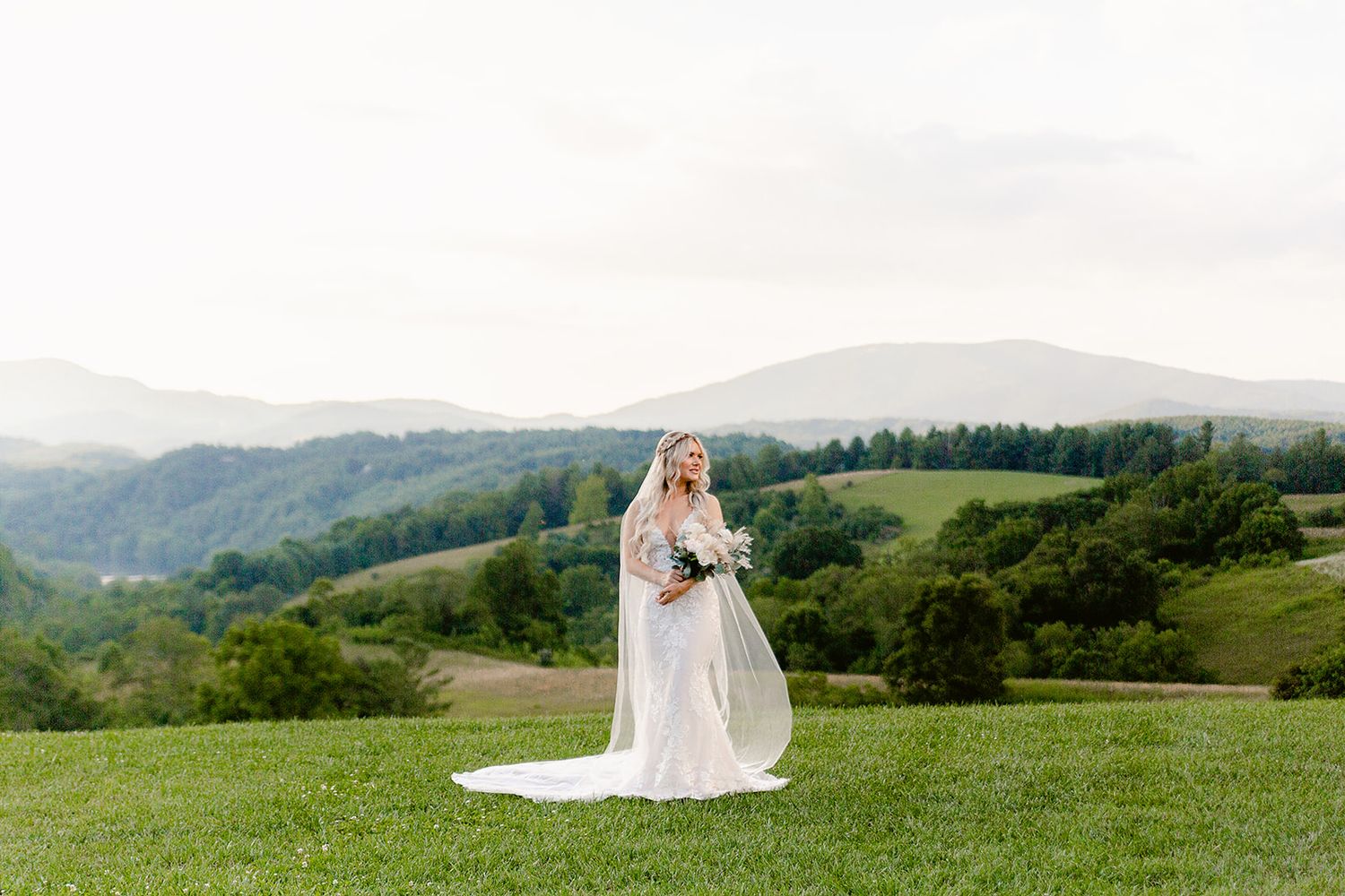 Scenic mountain view frames bride in flowing white gown and veil on grassy hillside.