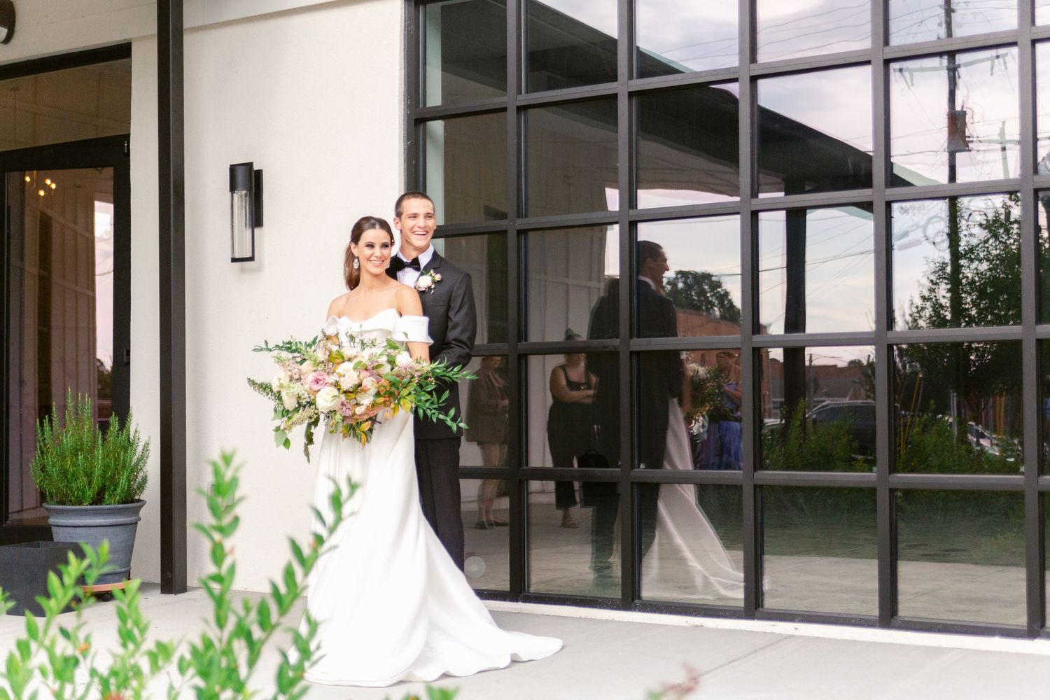 A couple poses in front of modern glass doors at an elegant venue entrance with a lush floral bouquet.