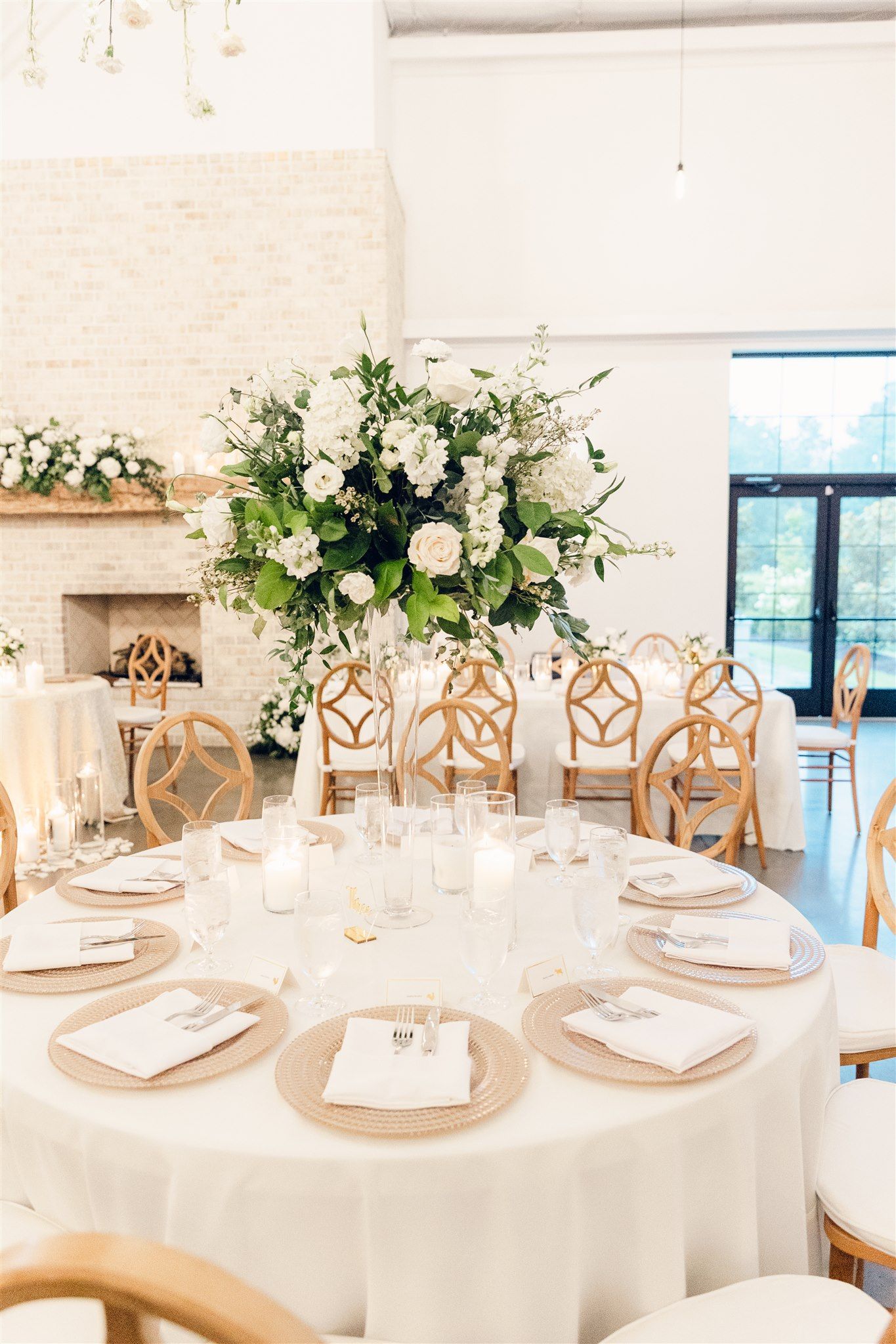 Elegant white table settings with gold accents and tall floral centerpieces in a bright modern reception venue.