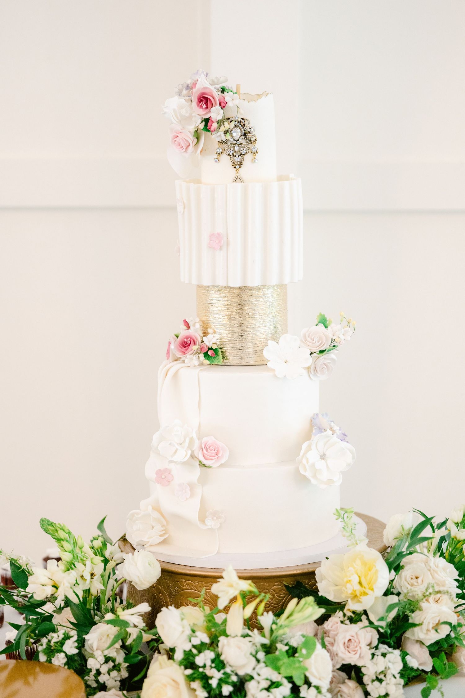 Elegant white wedding cake with pink roses and gold accents displayed on a floral-decorated dessert table.