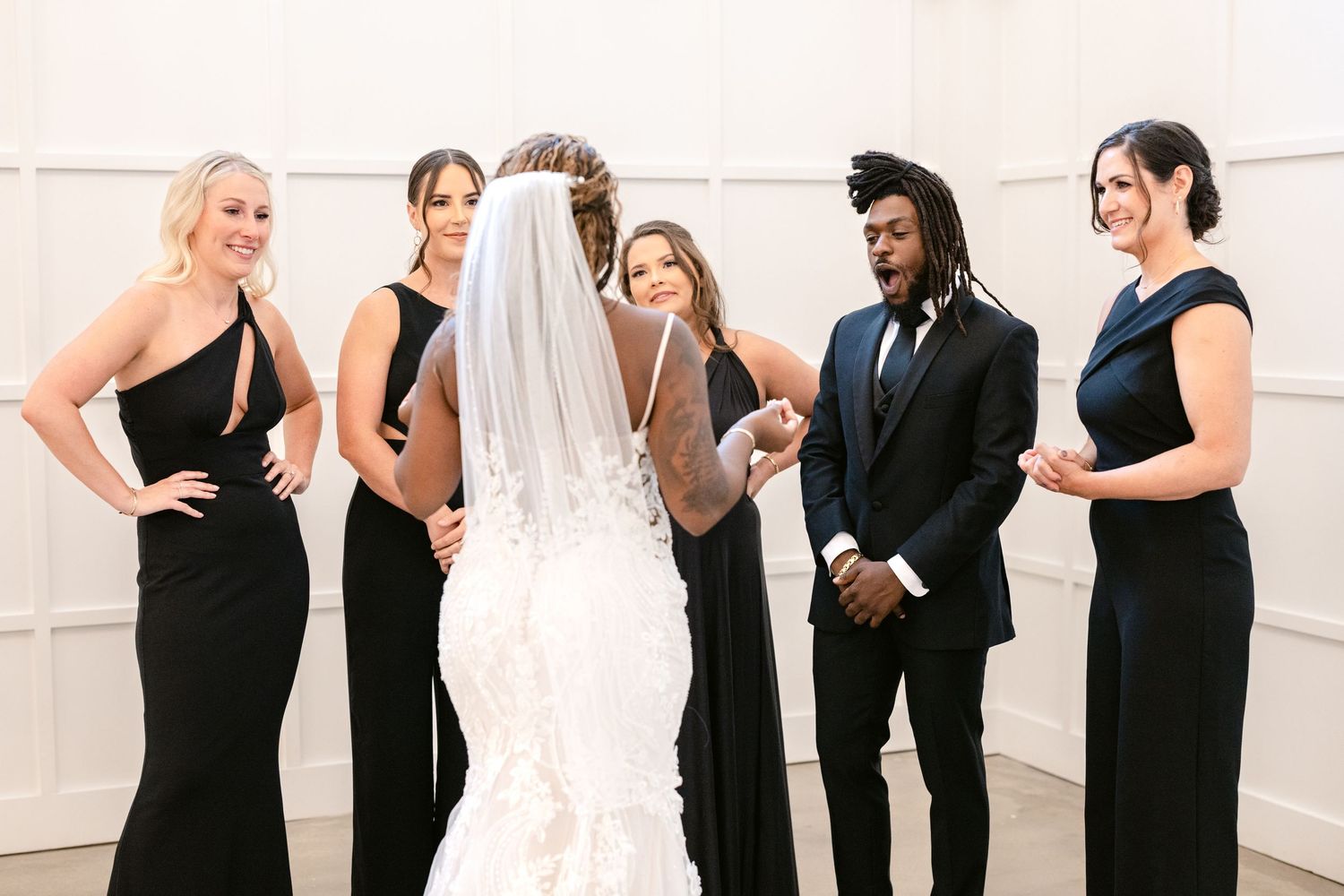 Wedding party wearing elegant black attire gathers around bride in white gown and veil at formal celebration.