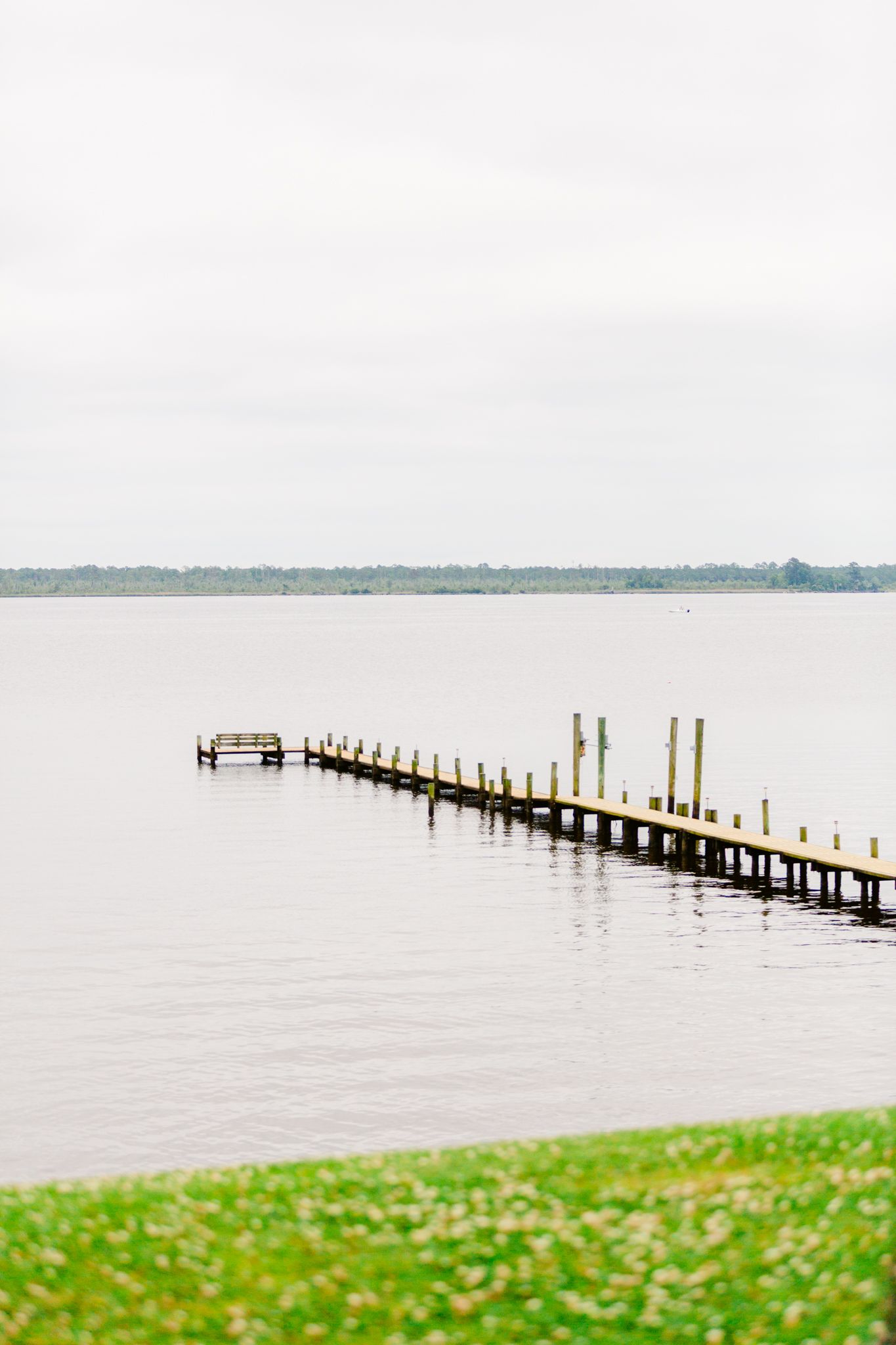 Wooden pier extends into a misty lake with green grass in the foreground on a cloudy day.