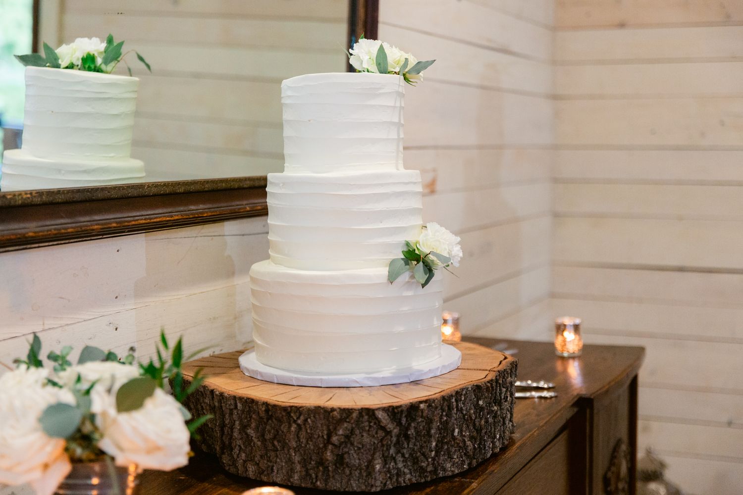 Elegant white tiered wedding cake displayed on rustic wood slice with delicate floral accents and candlelight.
