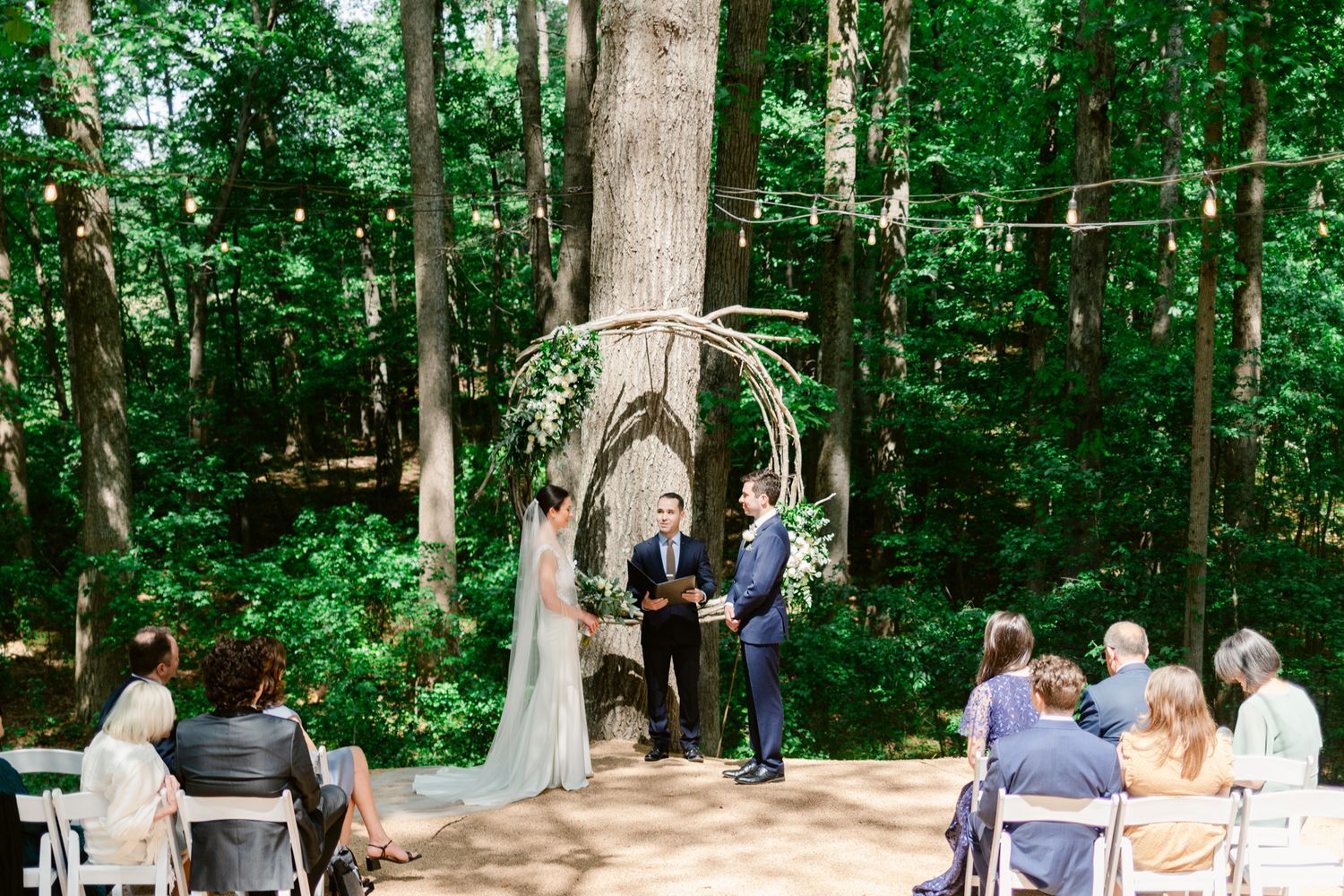 An outdoor wedding ceremony takes place in a lush wooded area with white chairs arranged in rows under tall trees.