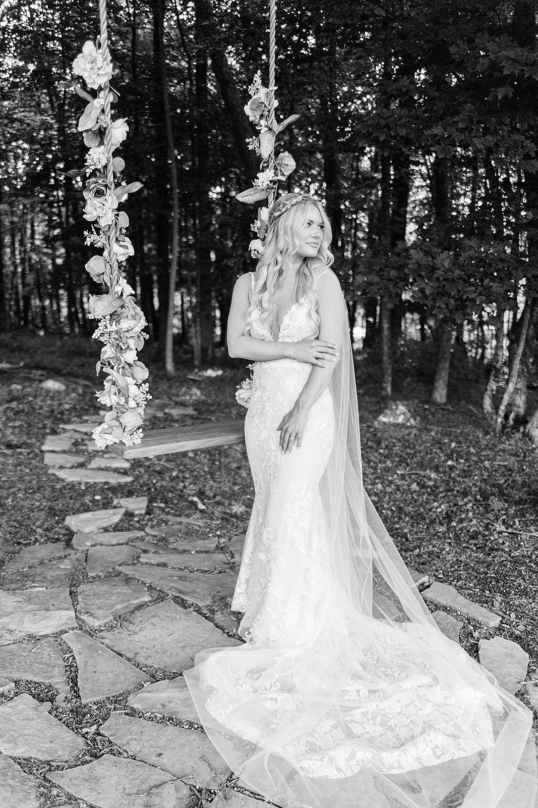 A bride in a flowing white lace wedding gown poses on stone steps in a wooded outdoor setting.