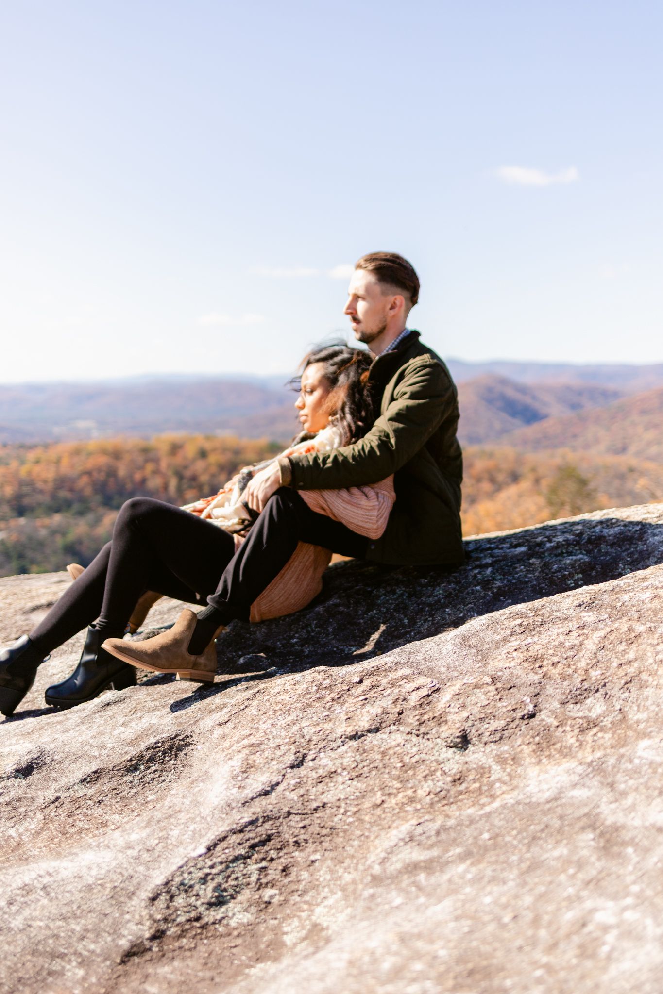 A couple relaxes together on a large rock formation overlooking a colorful fall mountain landscape.