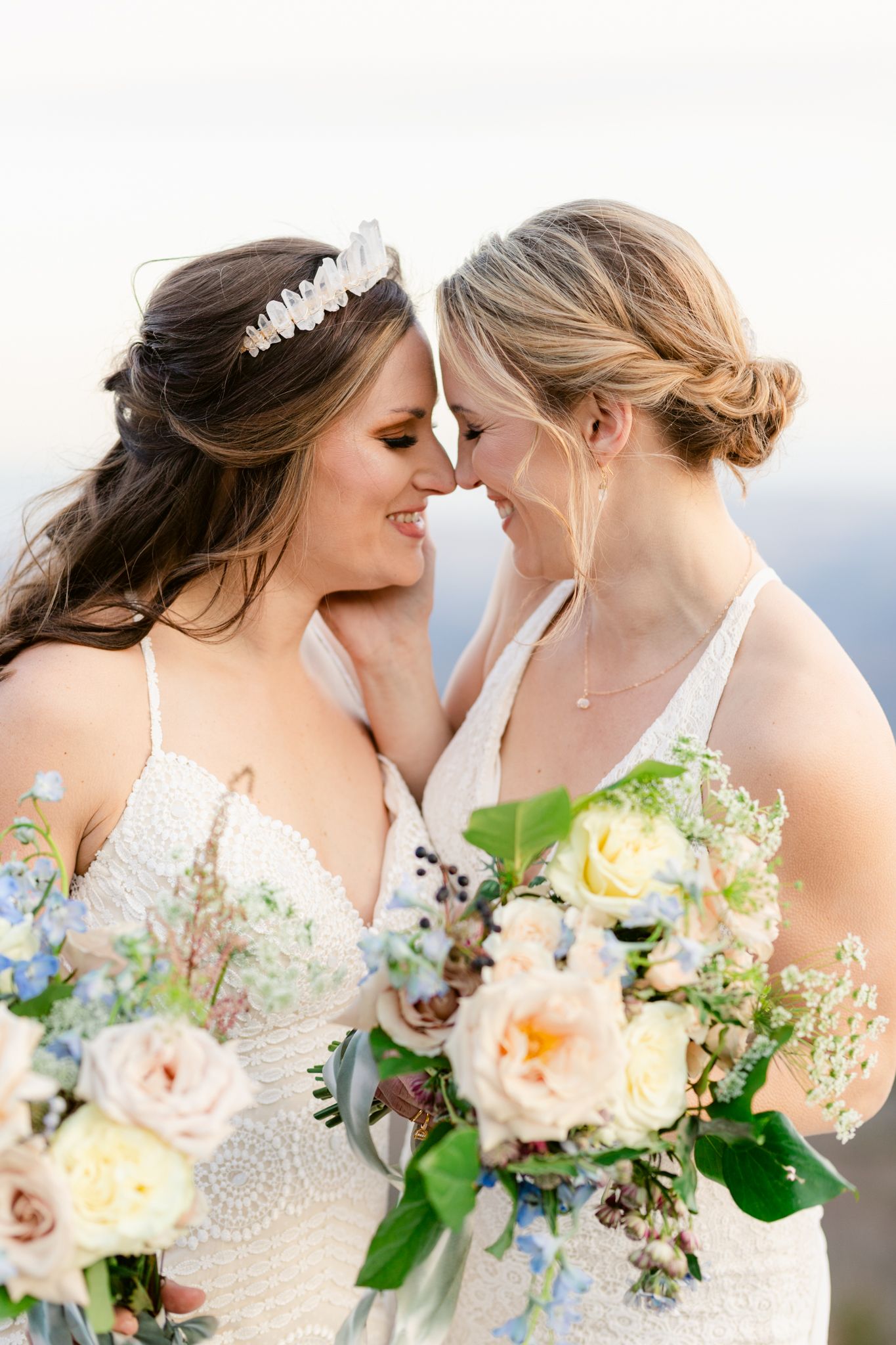 Two people in white dresses share a tender moment while holding delicate floral bouquets with peach and blue blooms.