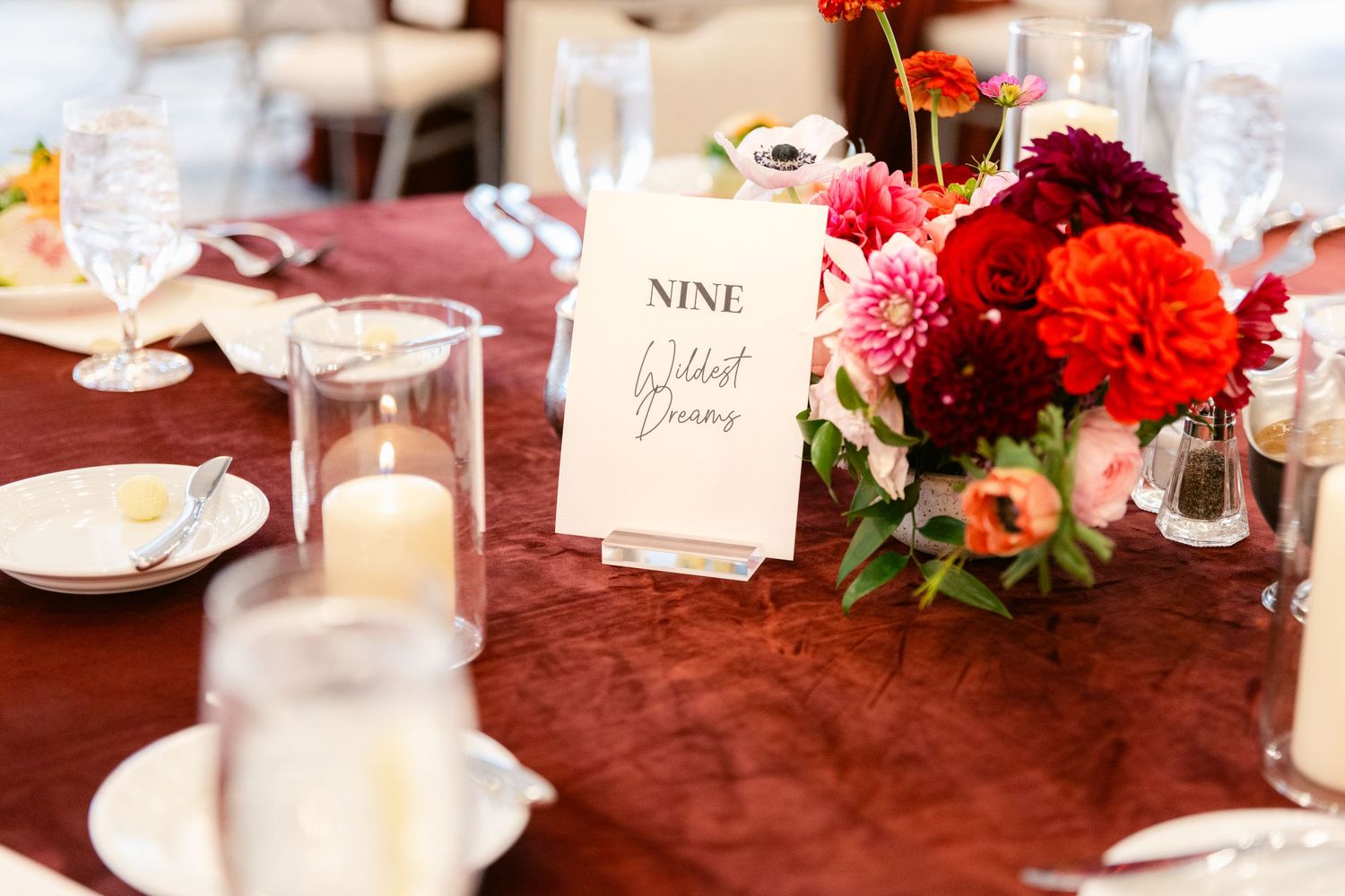 Elegant wedding reception table decorated with red and pink floral centerpieces, candles, and place settings on burgundy linens.