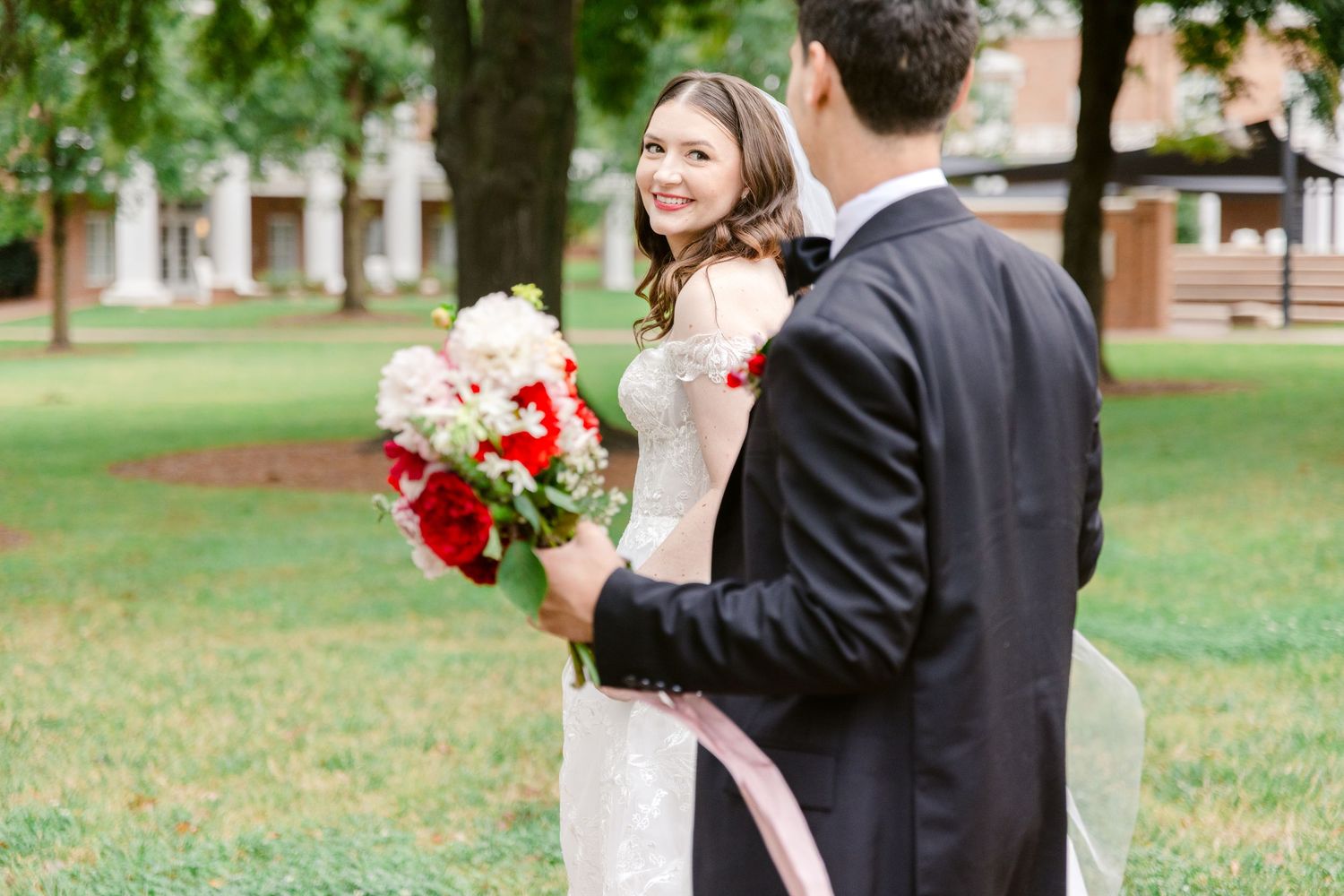 Bride in lace gown holds red and white bouquet, smiling at groom in black suit on green lawn.