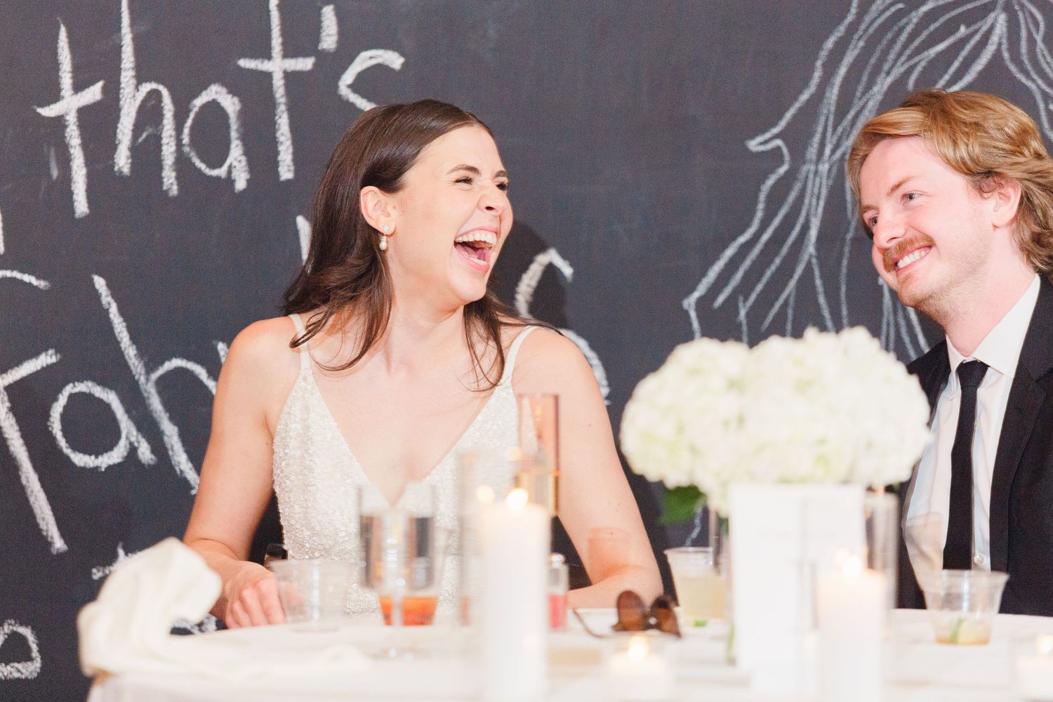 A couple laughing together at their wedding reception with a chalkboard wall backdrop and white floral centerpieces.