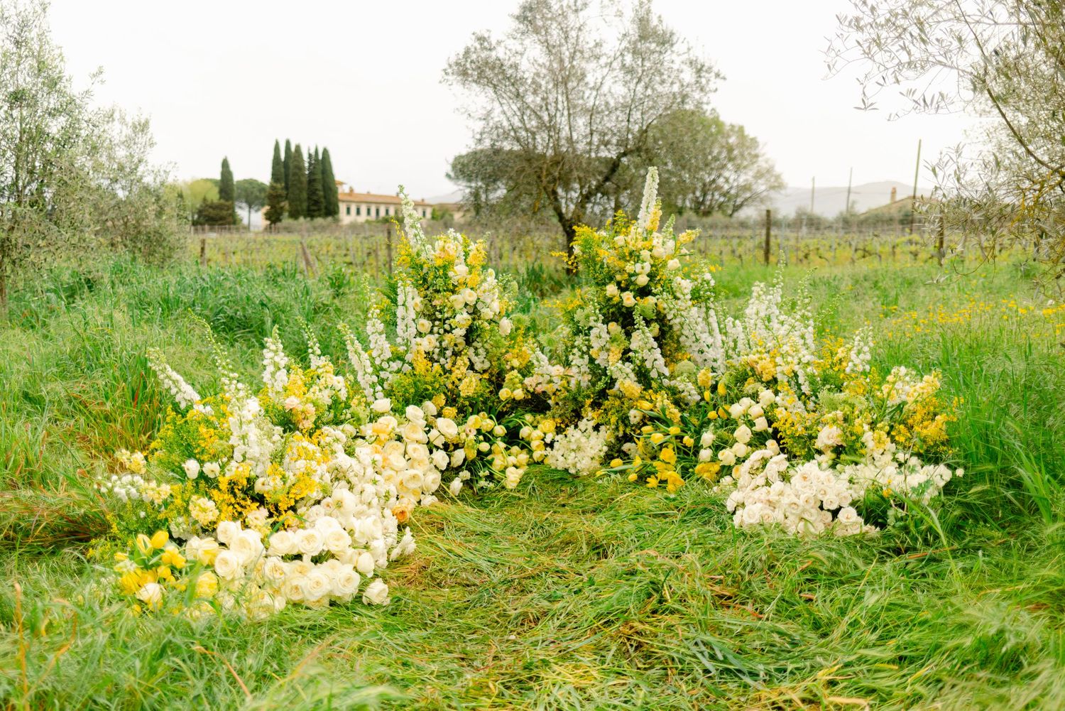 A curved floral arrangement with yellow and white flowers sits on grass in a garden landscape.