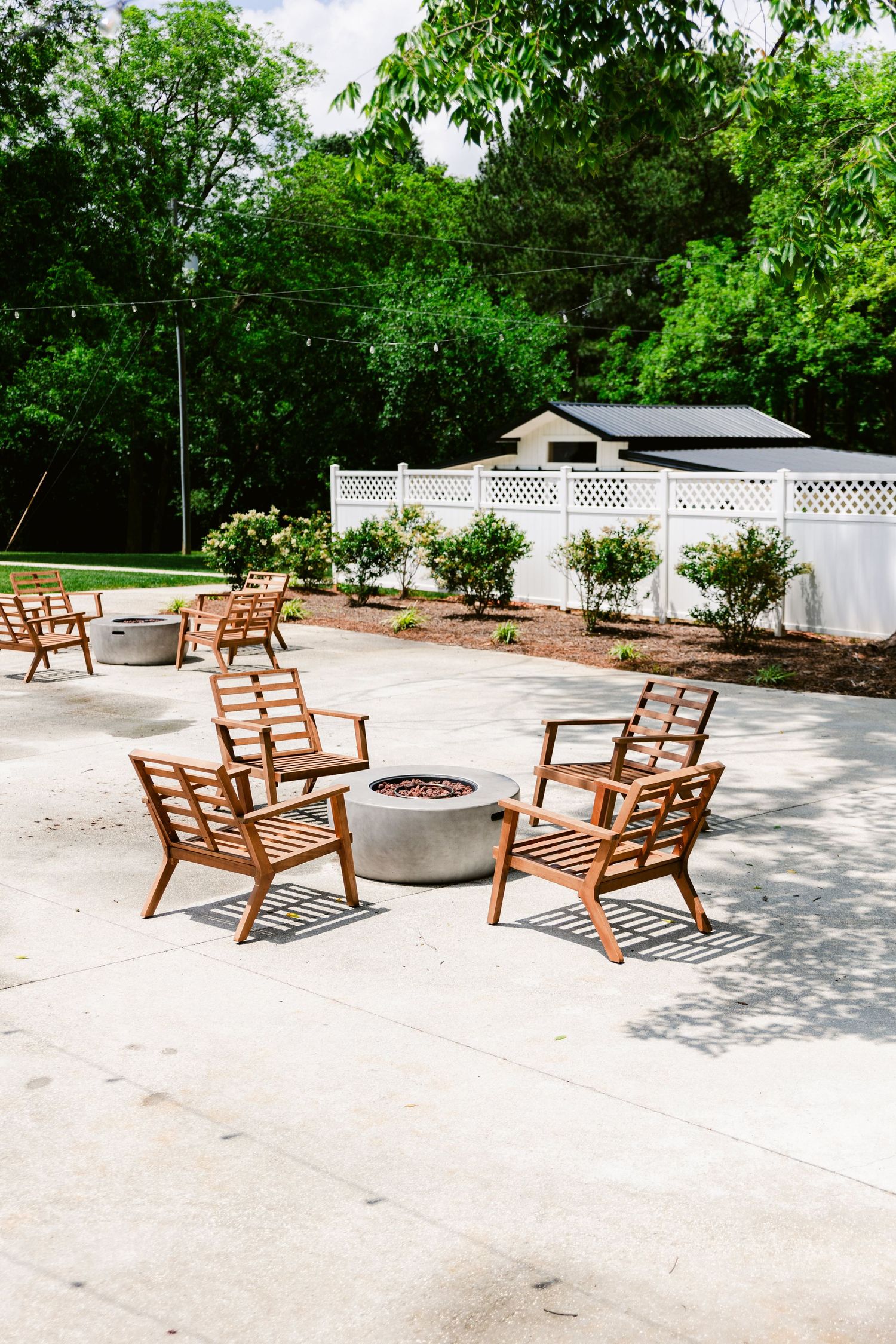 Wooden lounge chairs arranged around a fire pit on a concrete patio surrounded by greenery.