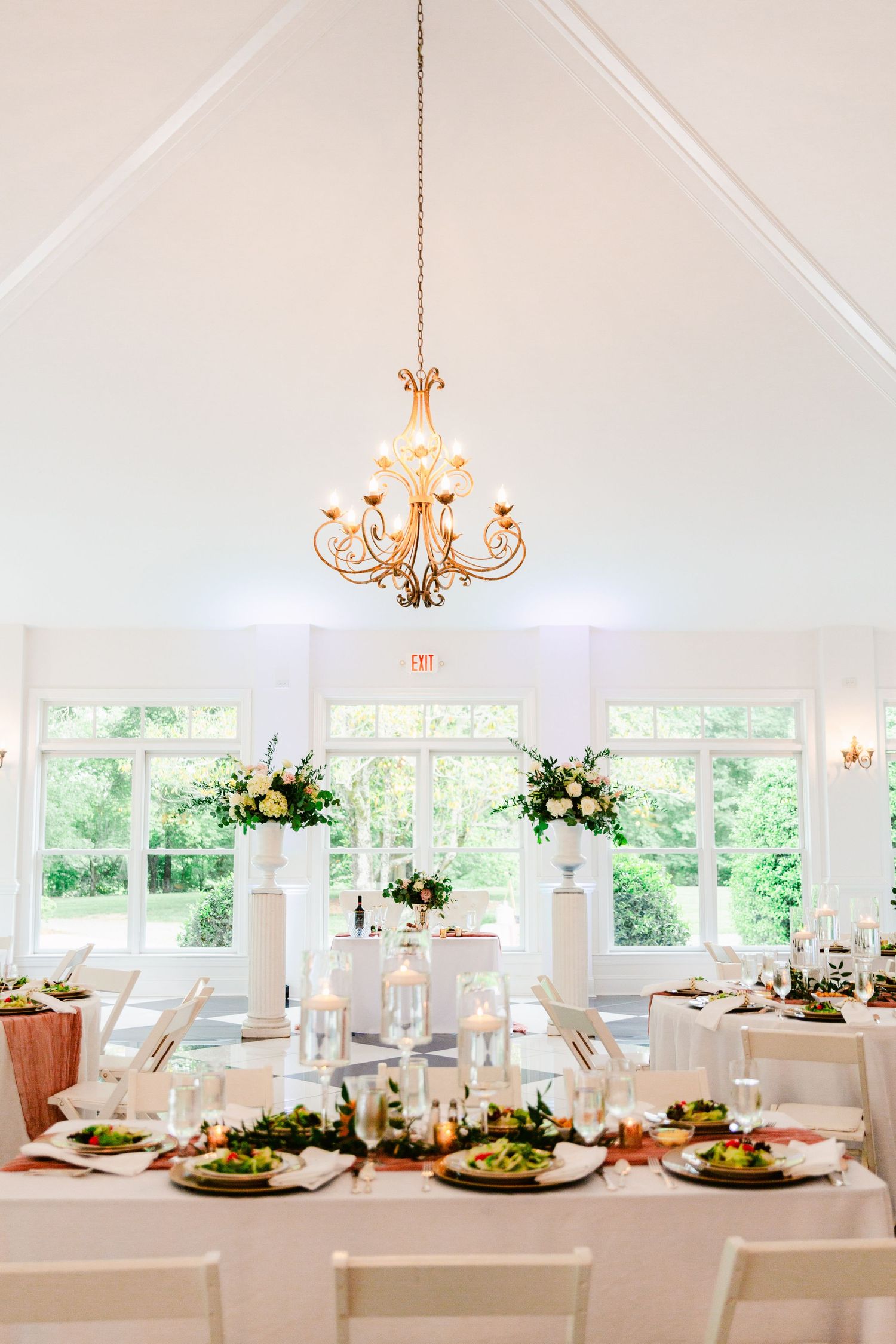 Bright white wedding venue interior with chandeliers and table settings arranged under a vaulted ceiling.