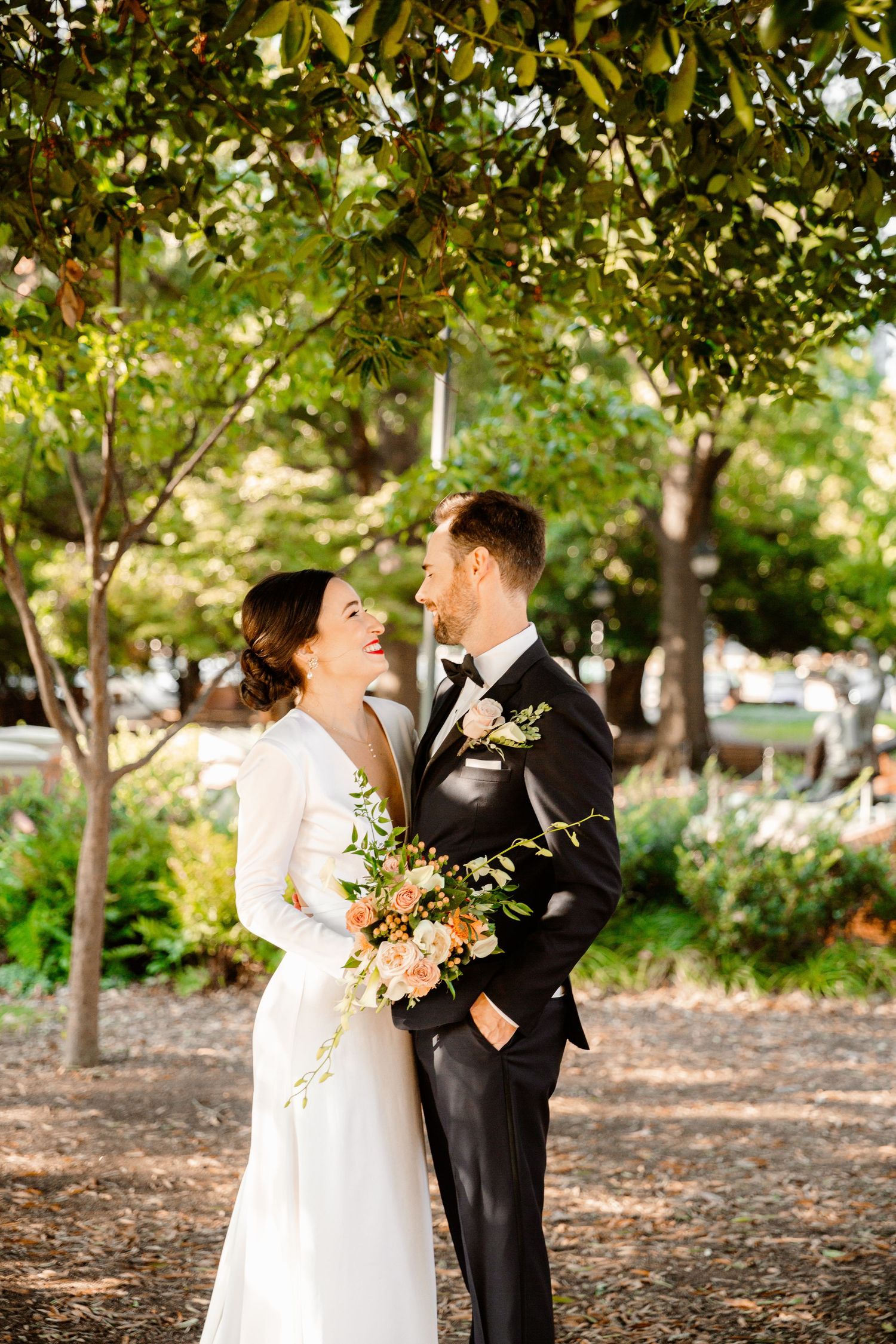 A wedding couple embraces outdoors among fall foliage while holding a peach-colored bouquet.