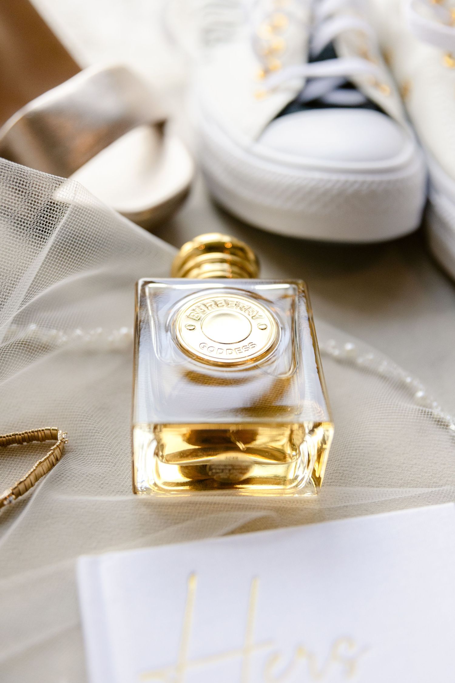 Elegant perfume bottle with gold cap displayed next to white sneakers and delicate tulle fabric.