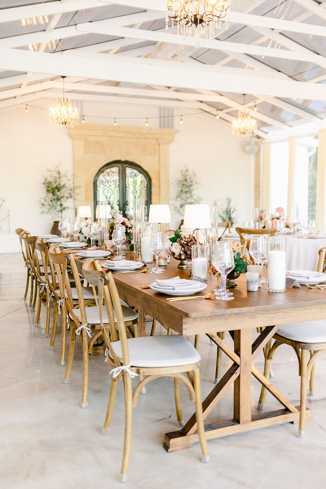 Elegant wedding reception venue with white walls, crystal chandeliers and wooden farm tables set for dining.