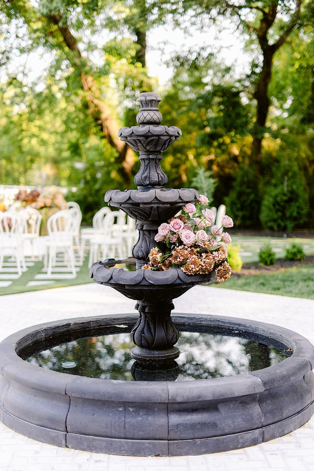 Elegant tiered fountain with pink floral arrangement in an outdoor wedding venue setting with white chairs in background.