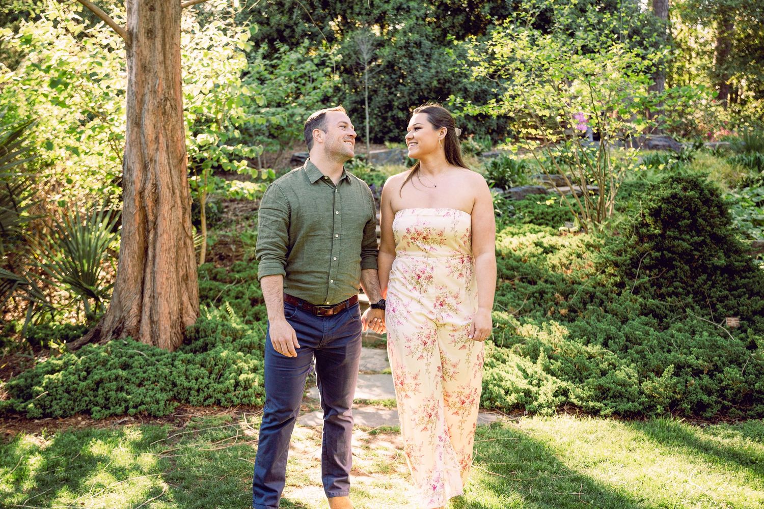A couple stands together on a sunny lawn surrounded by lush trees and greenery in a garden setting.