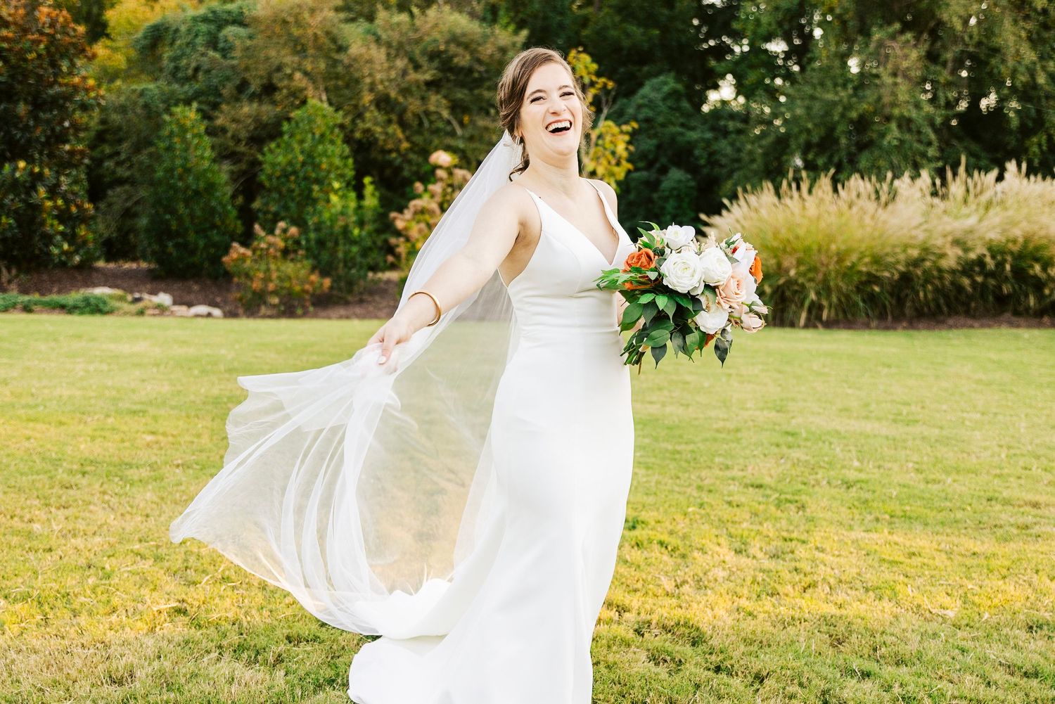 A bride in a flowing white dress twirls joyfully in a sunlit meadow during a summer wedding photoshoot.