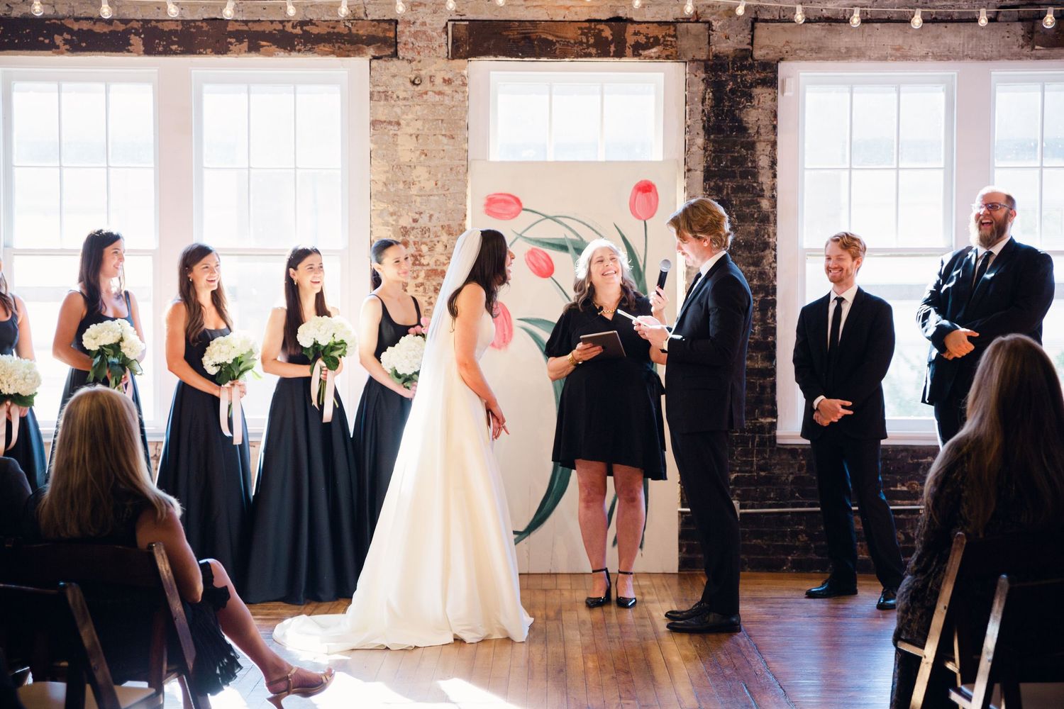 Wedding party members stand in a line during a ceremony in a rustic brick venue with large windows.
