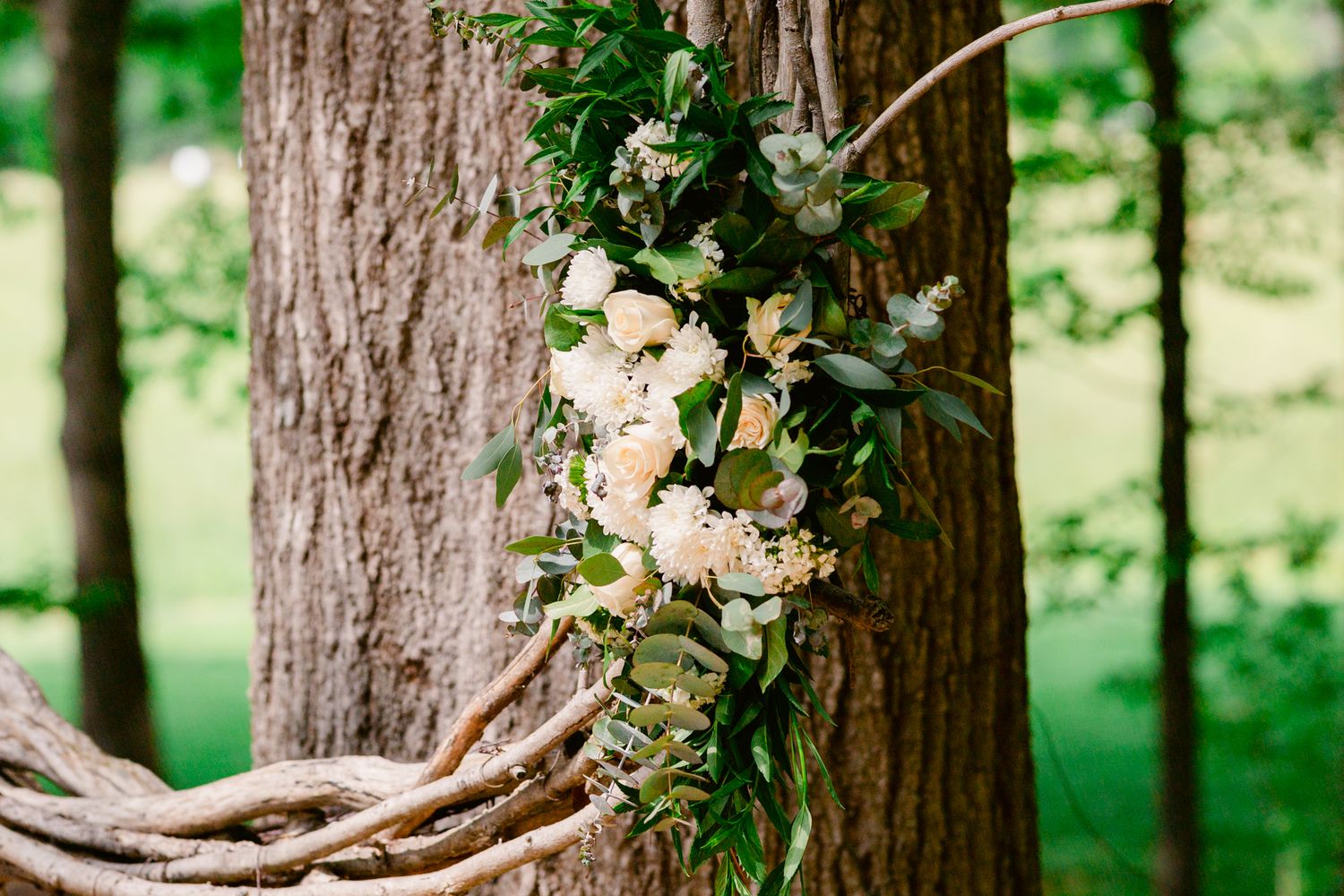 Delicate white floral arrangement with greenery wrapped around a rustic tree trunk in a forest setting.