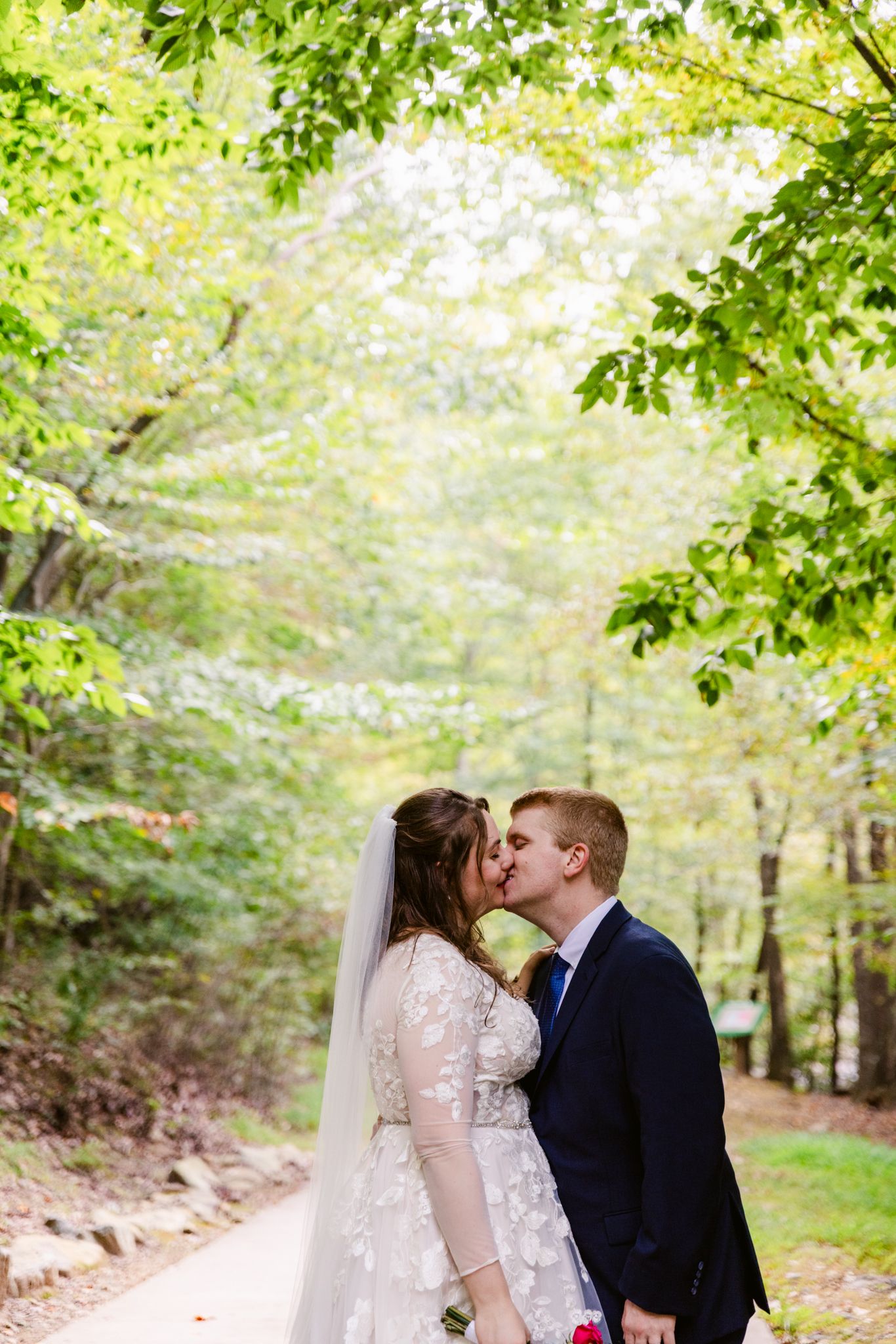 A romantic wedding couple shares a kiss on a tree-lined path surrounded by lush green foliage on their special day.
