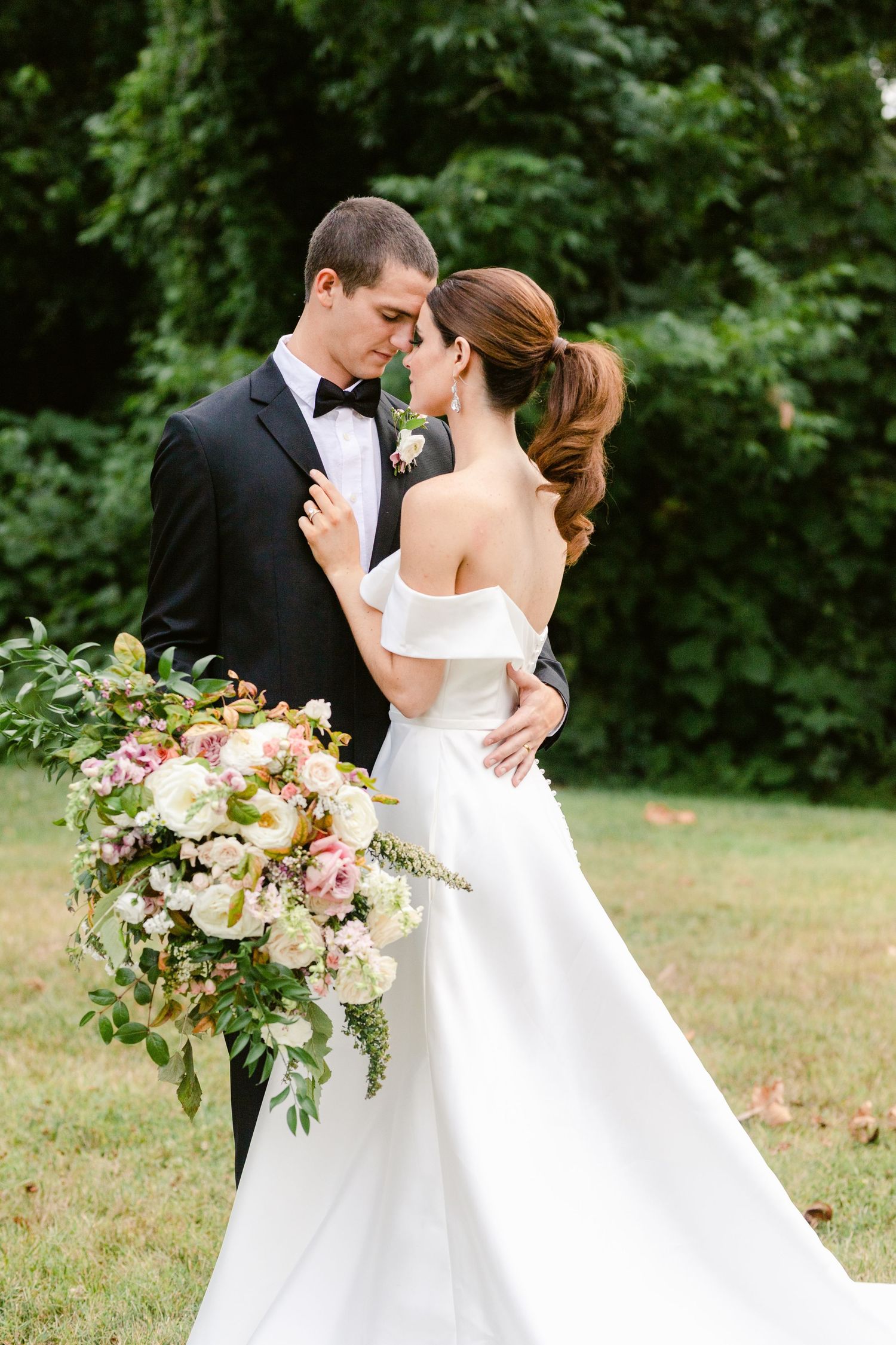 A romantic wedding couple embraces in a garden setting with a lush bouquet of pink and white flowers.