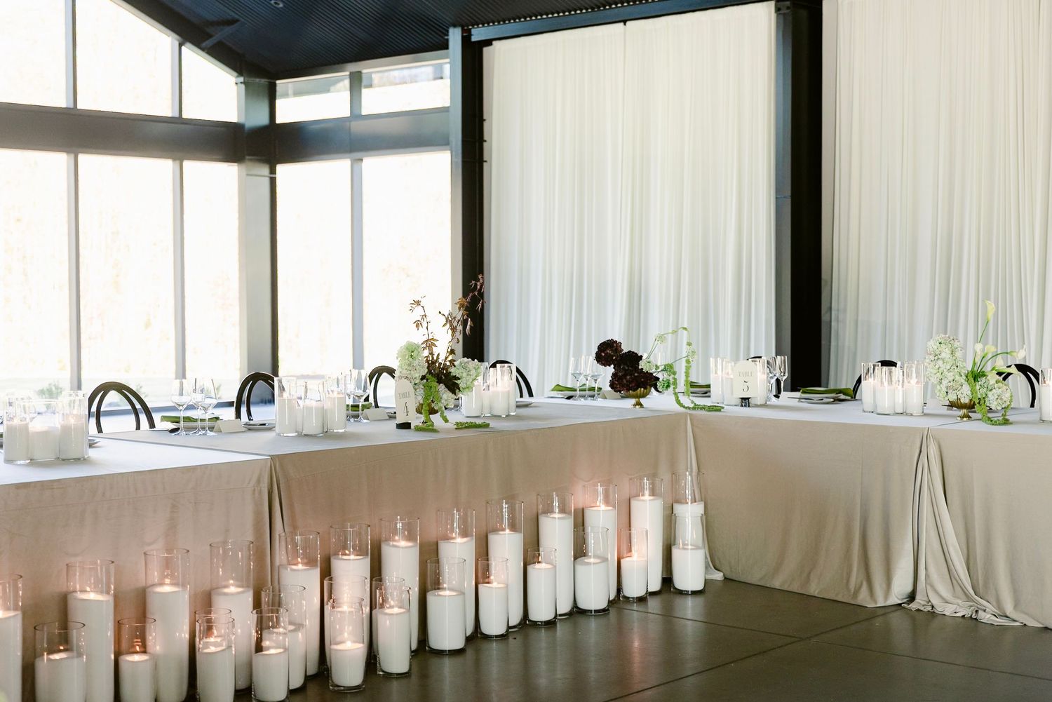 Long banquet table setup with white tablecloths and glowing candle centerpieces in a modern event space.