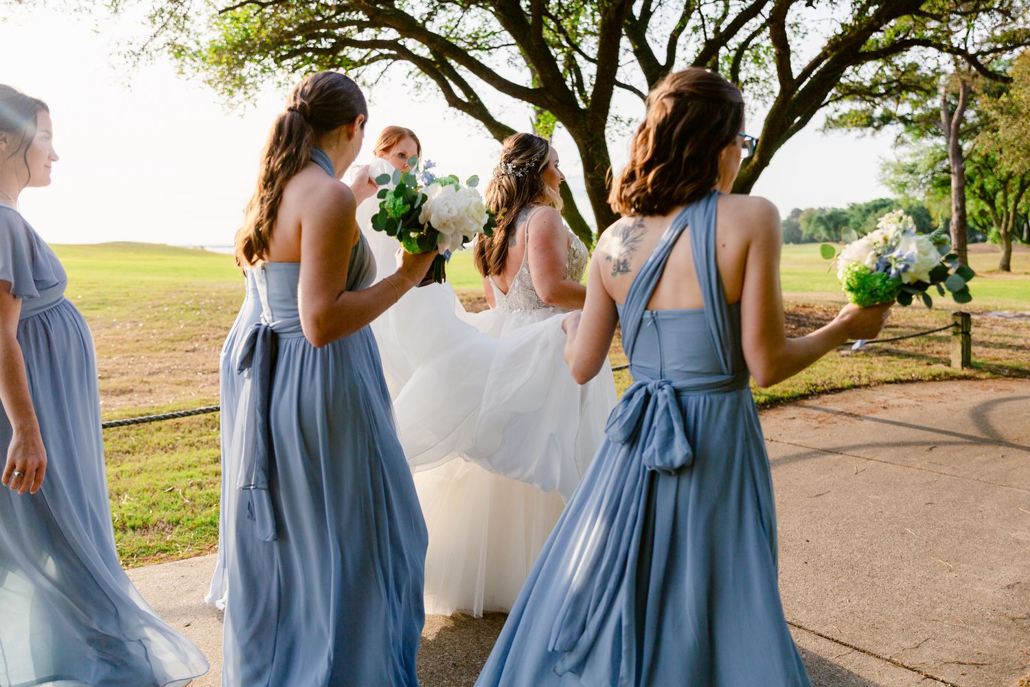 Bridesmaids in flowing dusty blue dresses gather with bride on sunny pathway during outdoor wedding celebration.
