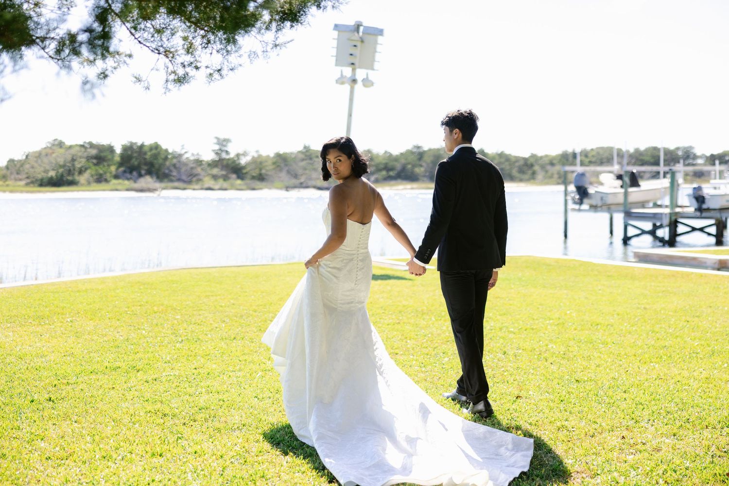 A couple stands together on a grassy waterfront lawn with a marina in the background.