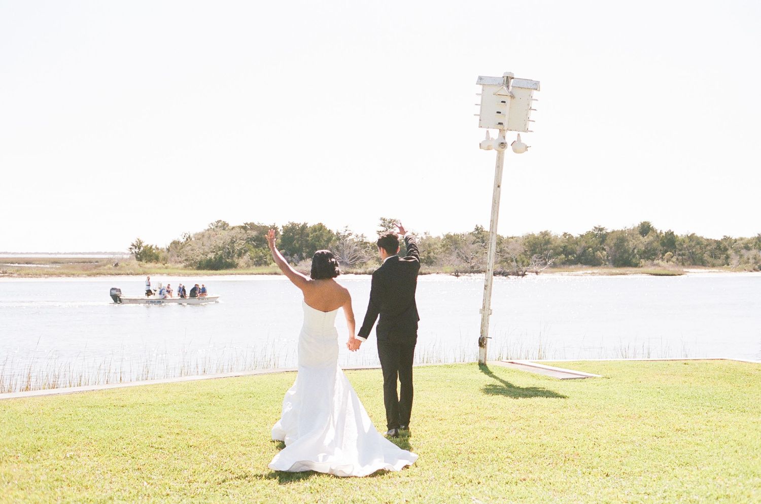 Couple in wedding attire walking along a scenic waterfront pathway with palm trees in the background.