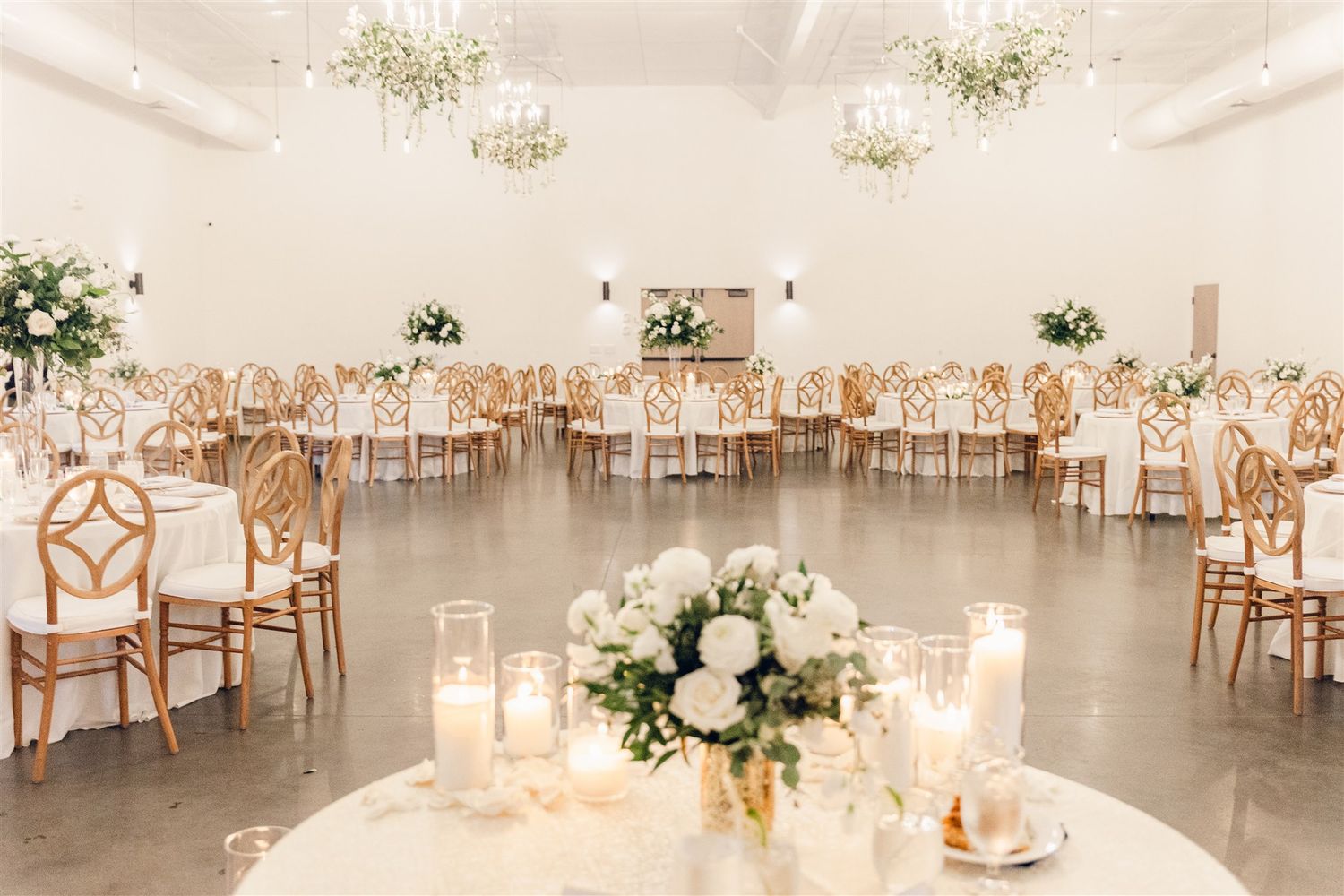 Elegant white wedding reception venue with wooden chairs, floral centerpieces and warm candlelight creating a romantic atmosphere.