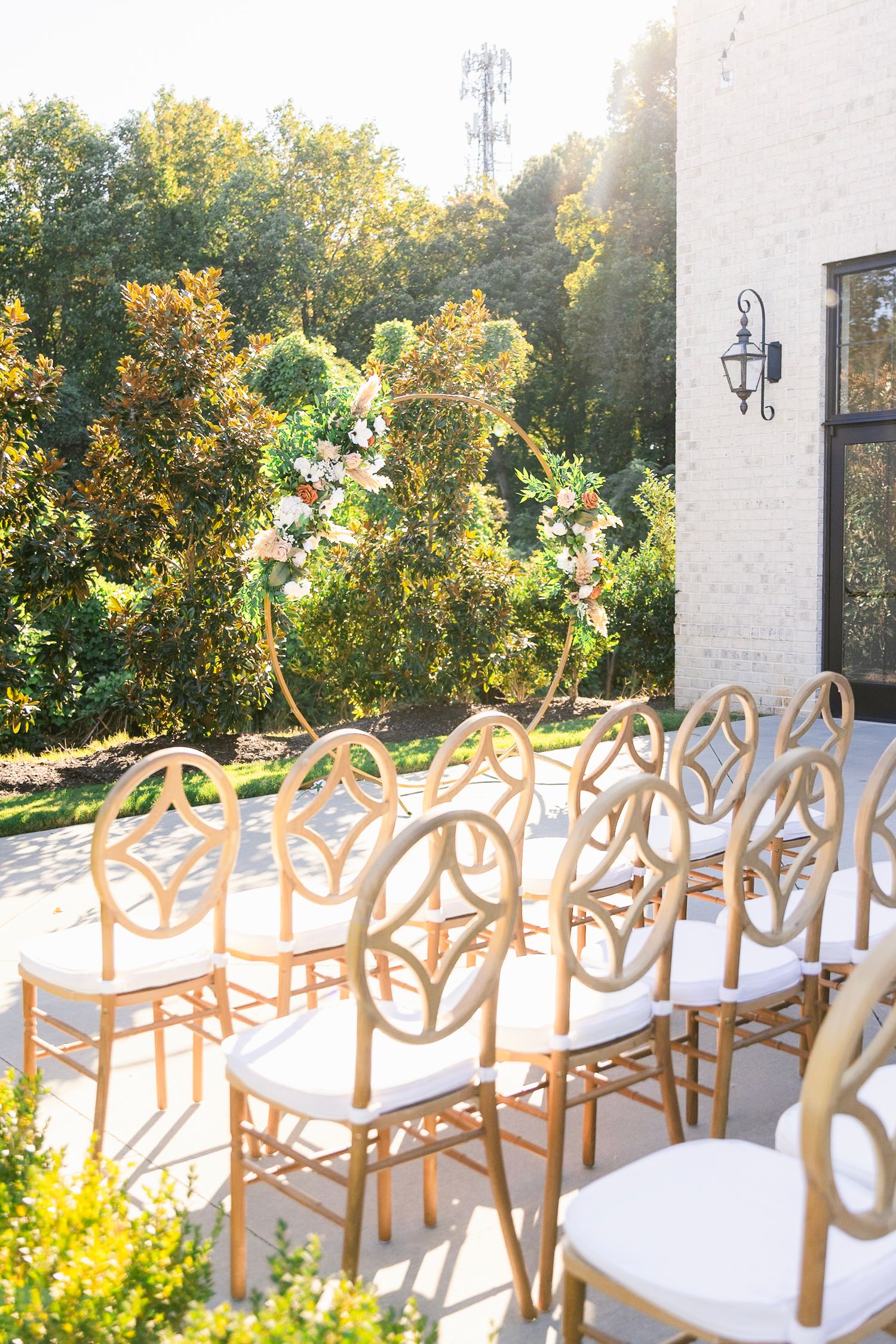 Elegant white and gold chairs arranged for an outdoor wedding ceremony with floral arch and sunlit garden backdrop.