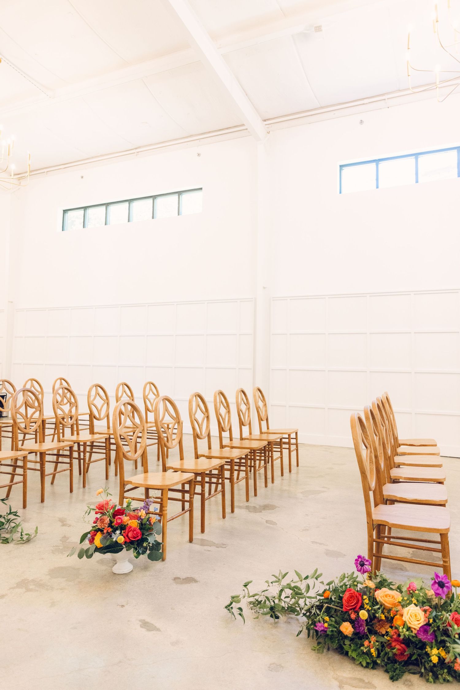Rows of wooden bentwood chairs arranged in a bright white room with colorful floral arrangements on the floor.