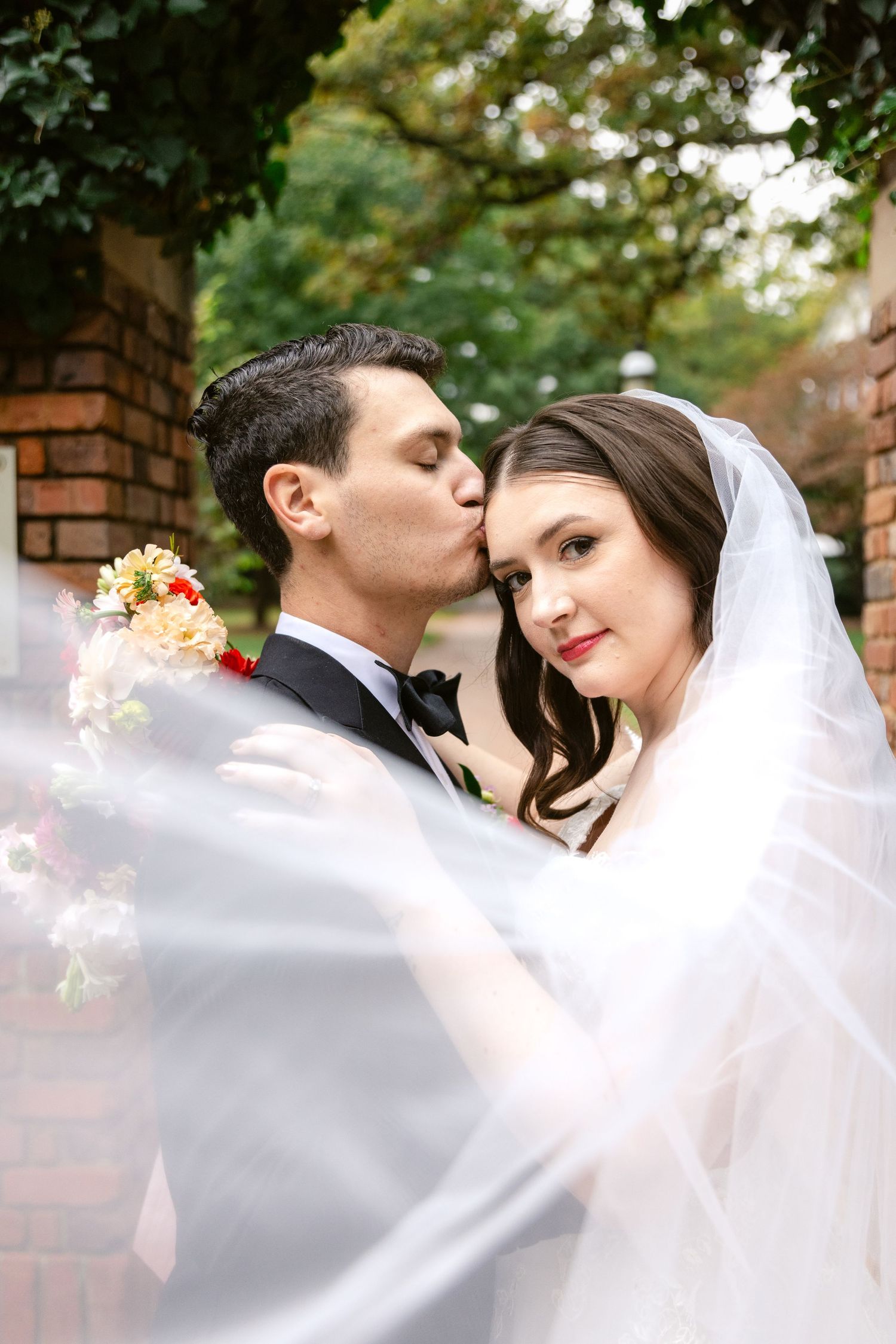 A romantic wedding moment captured through flowing white veil on a brick pathway.