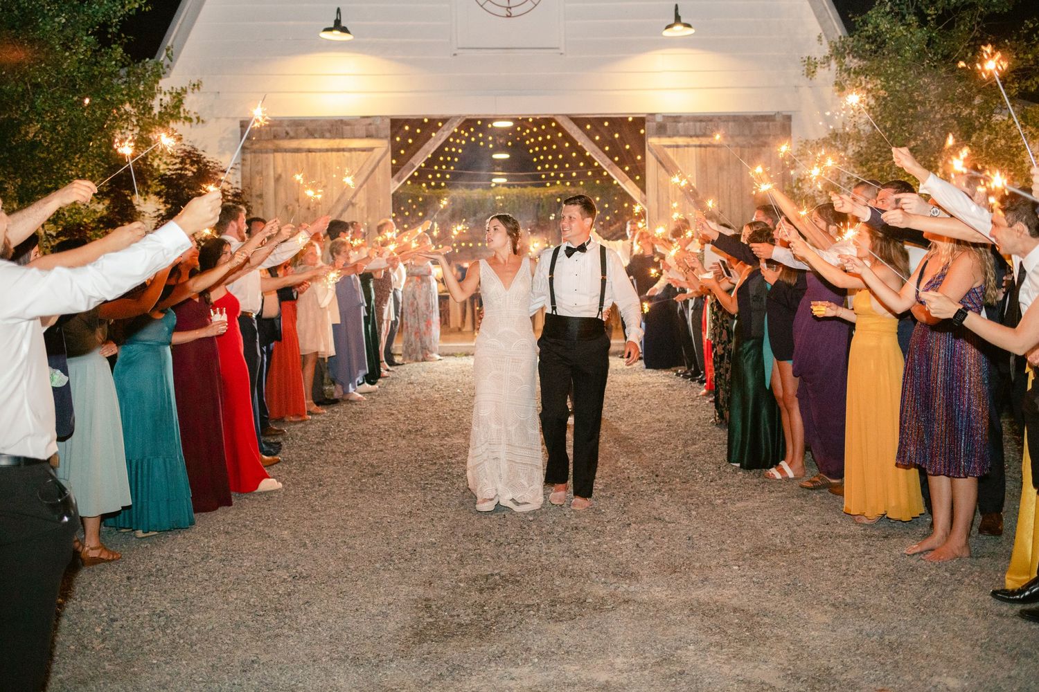 Newlyweds walk through sparkler exit tunnel as guests in colorful dresses line the path.