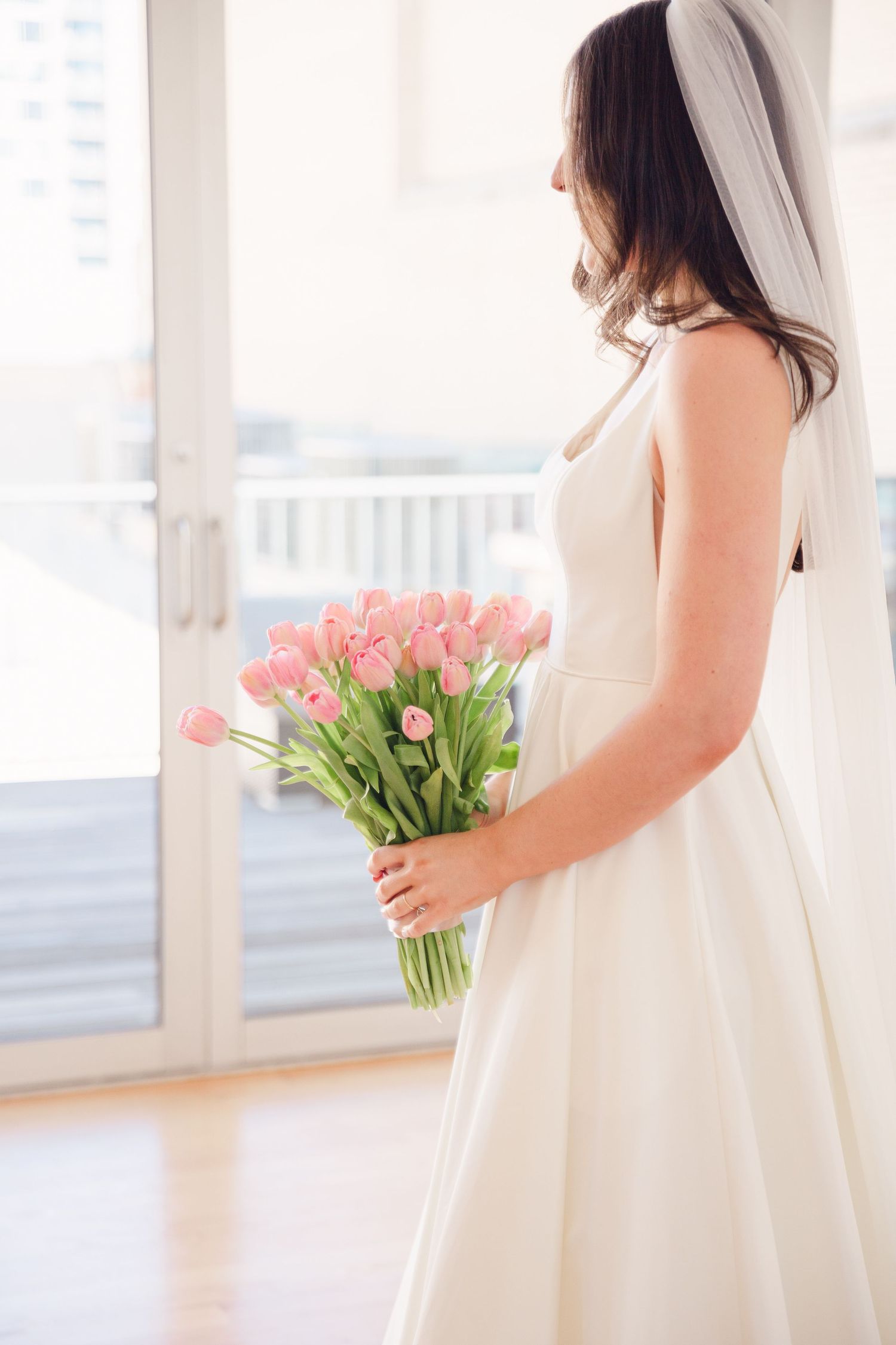 A person in a white wedding dress holds a bouquet of pink tulips while standing near a window.