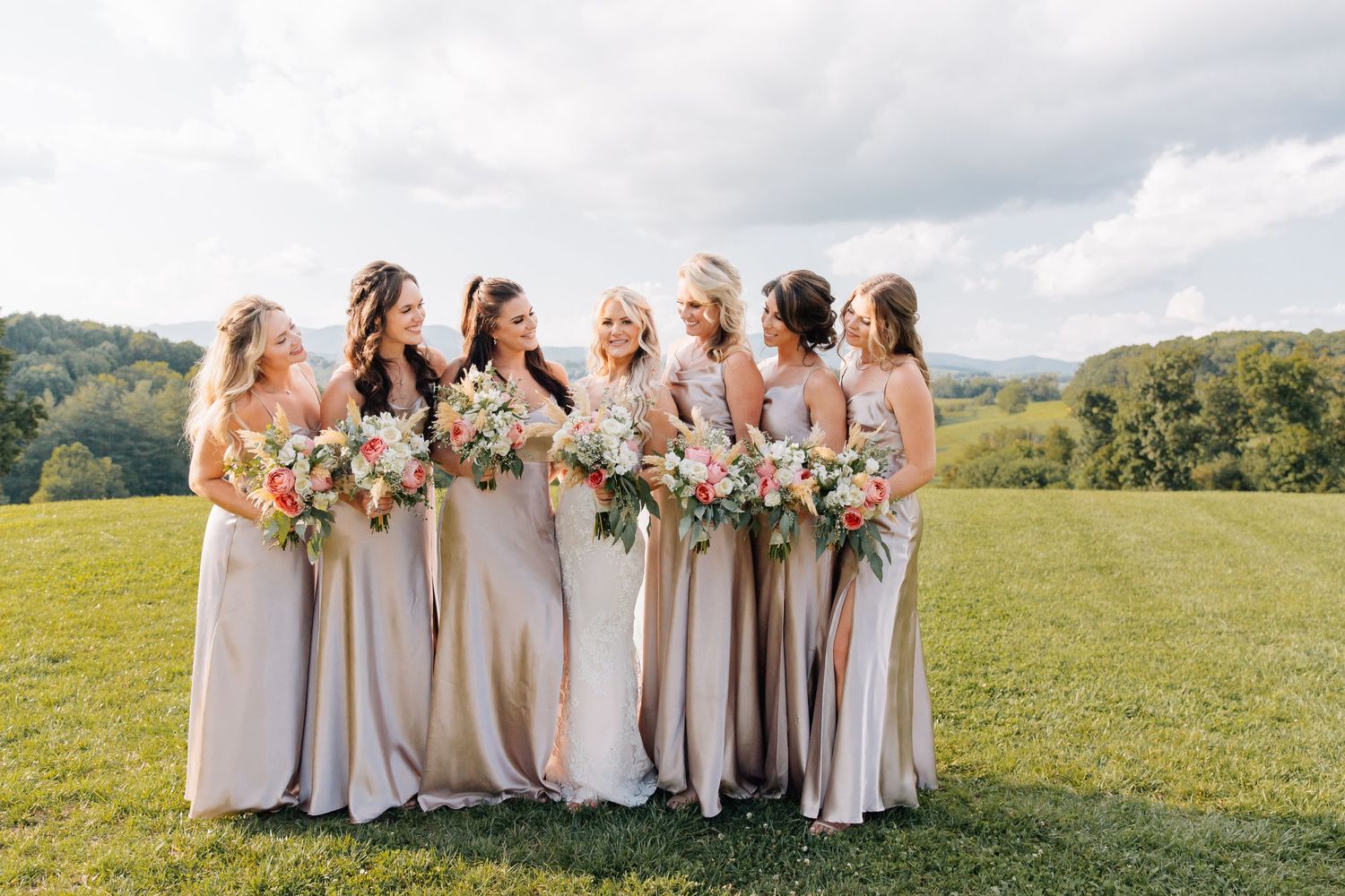 Bridesmaids in champagne dresses hold bouquets while standing in line on grassy hillside.