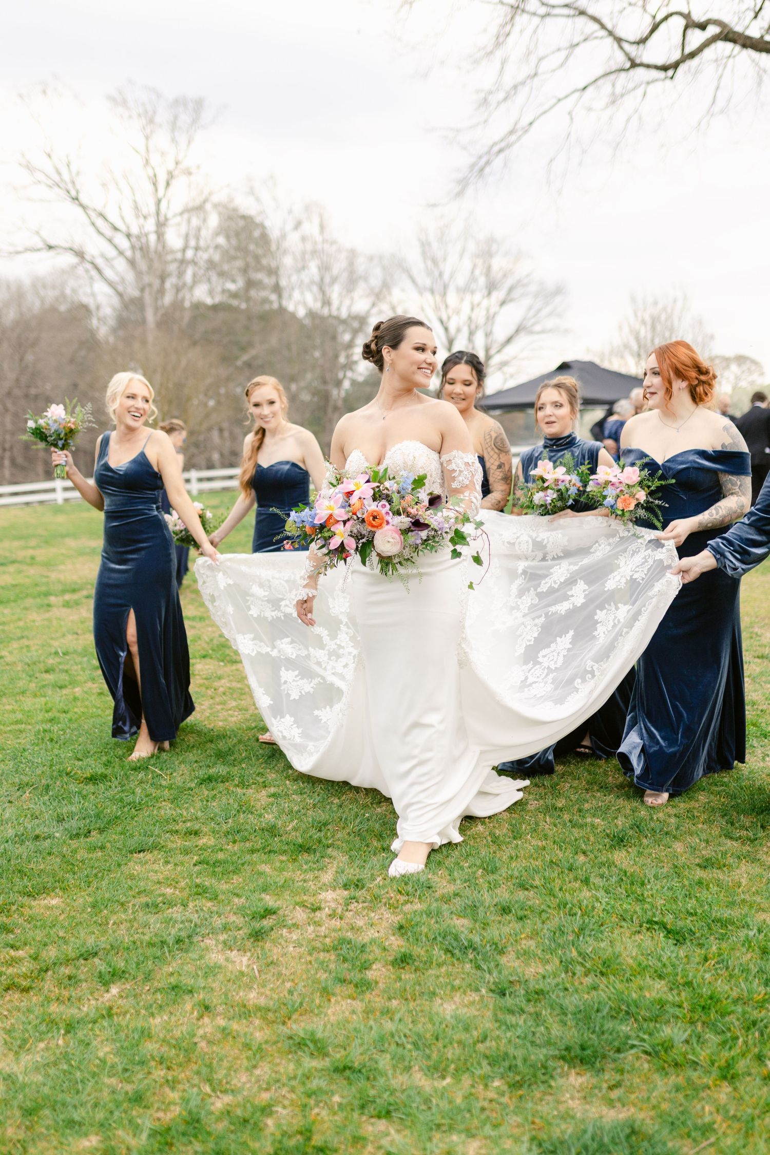 Wedding party in navy blue dresses help with the bride's flowing white gown on a grassy lawn.