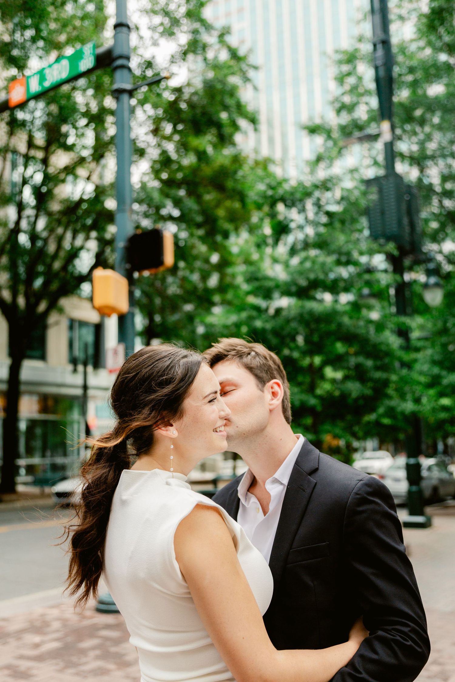 A couple sharing an intimate moment on a tree-lined urban street corner with traffic signals visible.