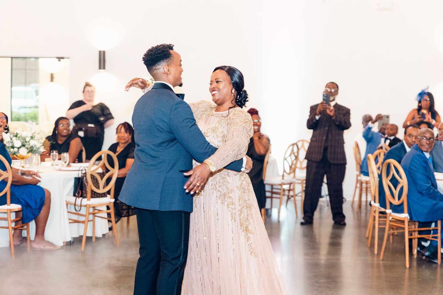 A couple shares their first dance at a wedding reception while guests watch from gold chairs in an elegant venue.