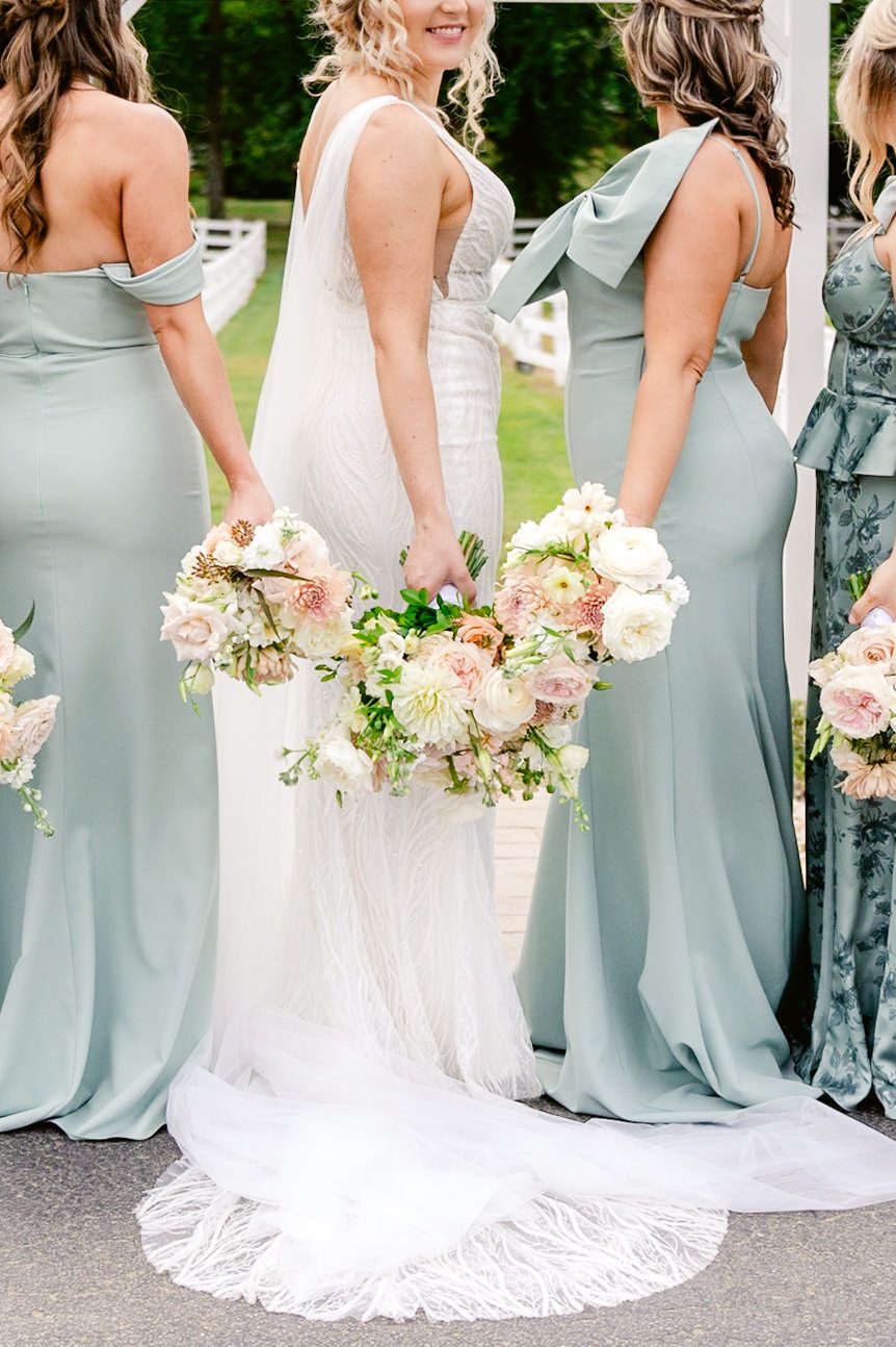 Bridesmaids in sage green dresses holding pink and white bouquets stand beside bride in flowing white wedding gown.