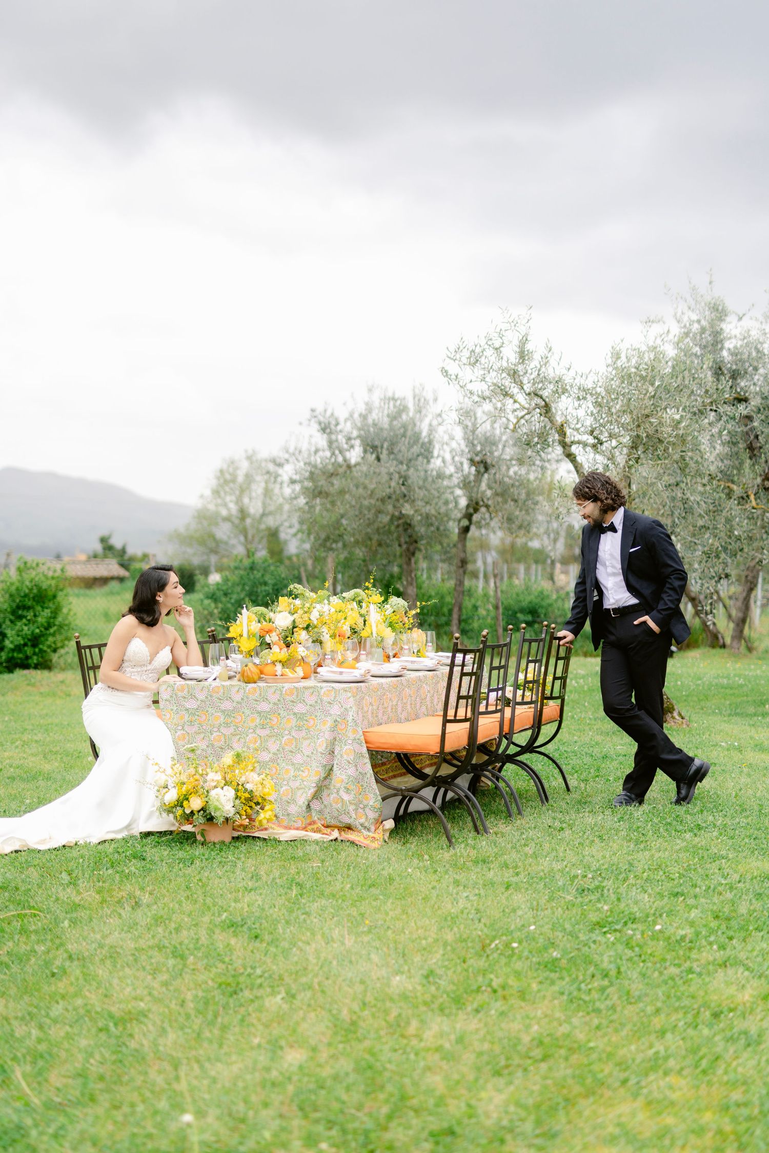 An elegant outdoor wedding table setting with yellow floral decor and iron chairs in a Tuscan olive grove.