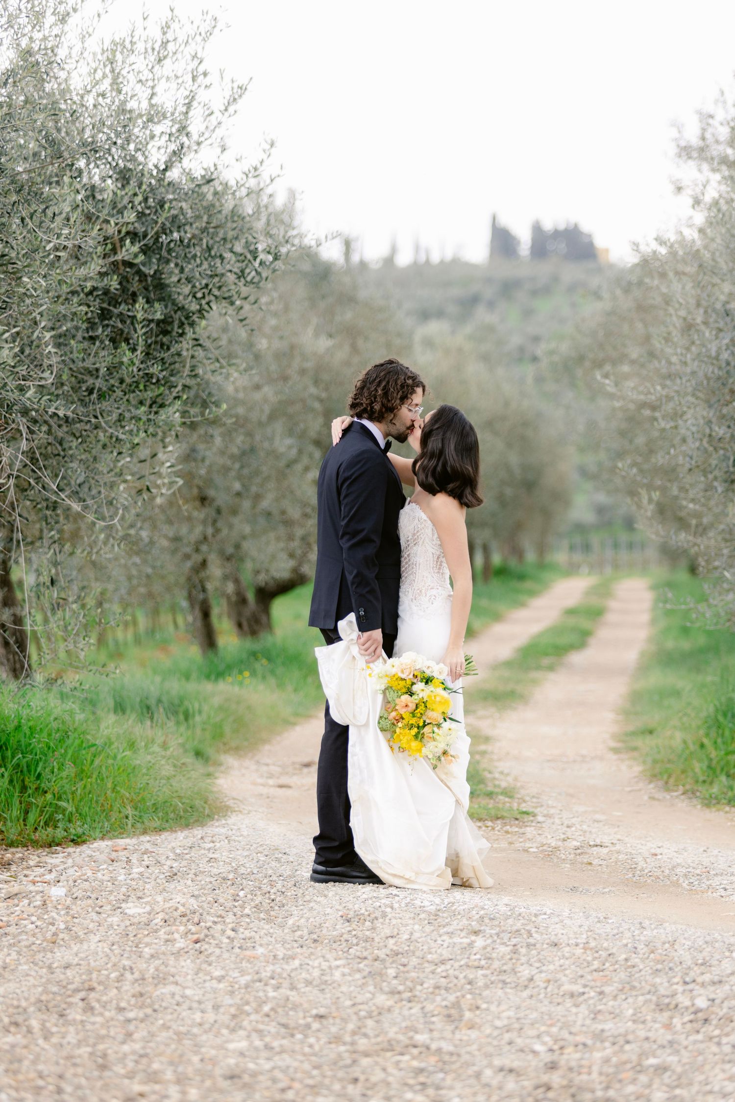 A romantic wedding moment captured on a rustic path lined with olive trees in the Italian countryside.