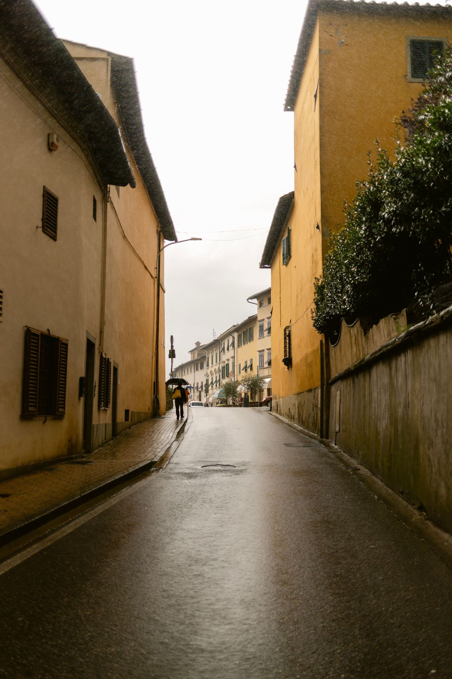 A lone figure with an umbrella walks down a rainy cobblestone street between yellow buildings in an Italian town.