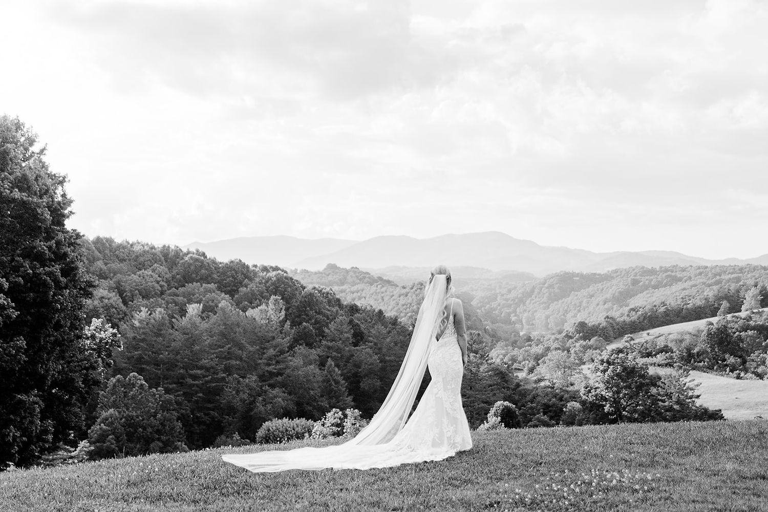 A long white wedding dress train flows across a grassy hilltop landscape with mountains in the background.