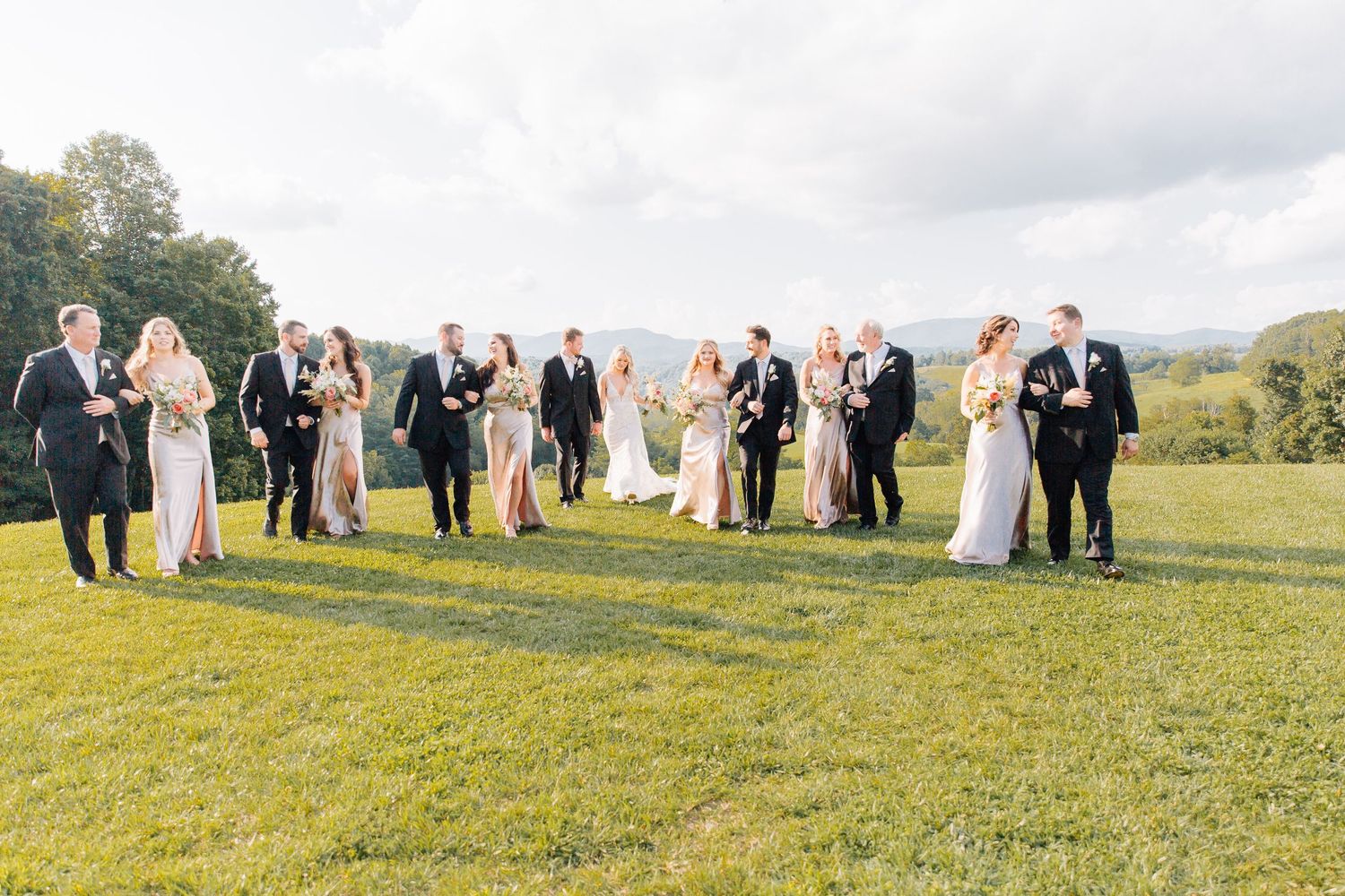 Wedding party members stand in a line formation on a grassy hillside during a sunny outdoor ceremony.