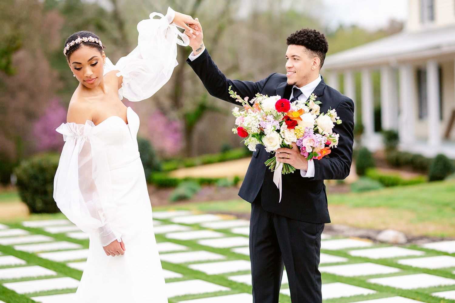A couple in wedding attire playfully interacts in a garden setting with white geometric paving stones and spring flowers.
