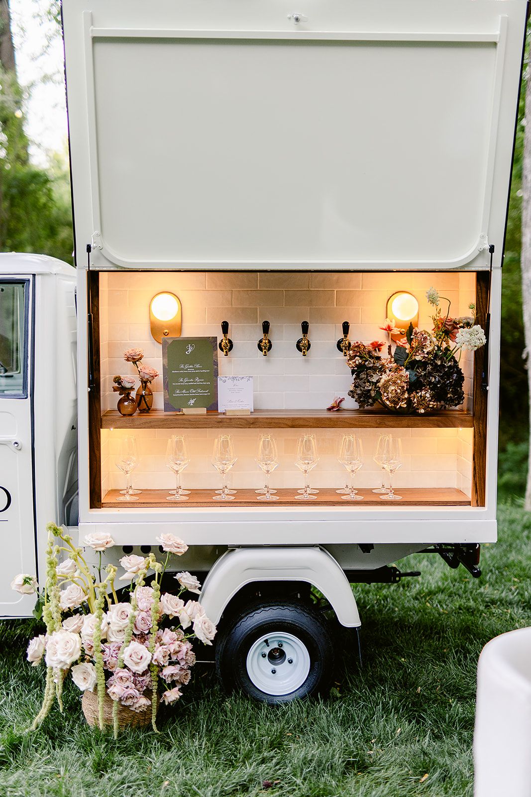 Vintage white mobile bar cart with illuminated shelves displaying glassware and floral arrangements at an outdoor event.